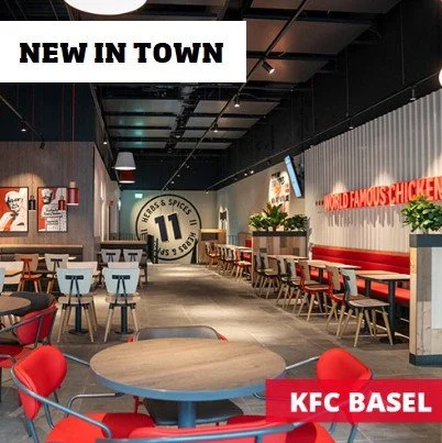 Innenansicht eines Fast-Food-Restaurants mit Tischen und Stühlen, einem Schild mit der Nummer 11, und einem KFC-Logo mit der Aufschrift 'KFC BASEL'.