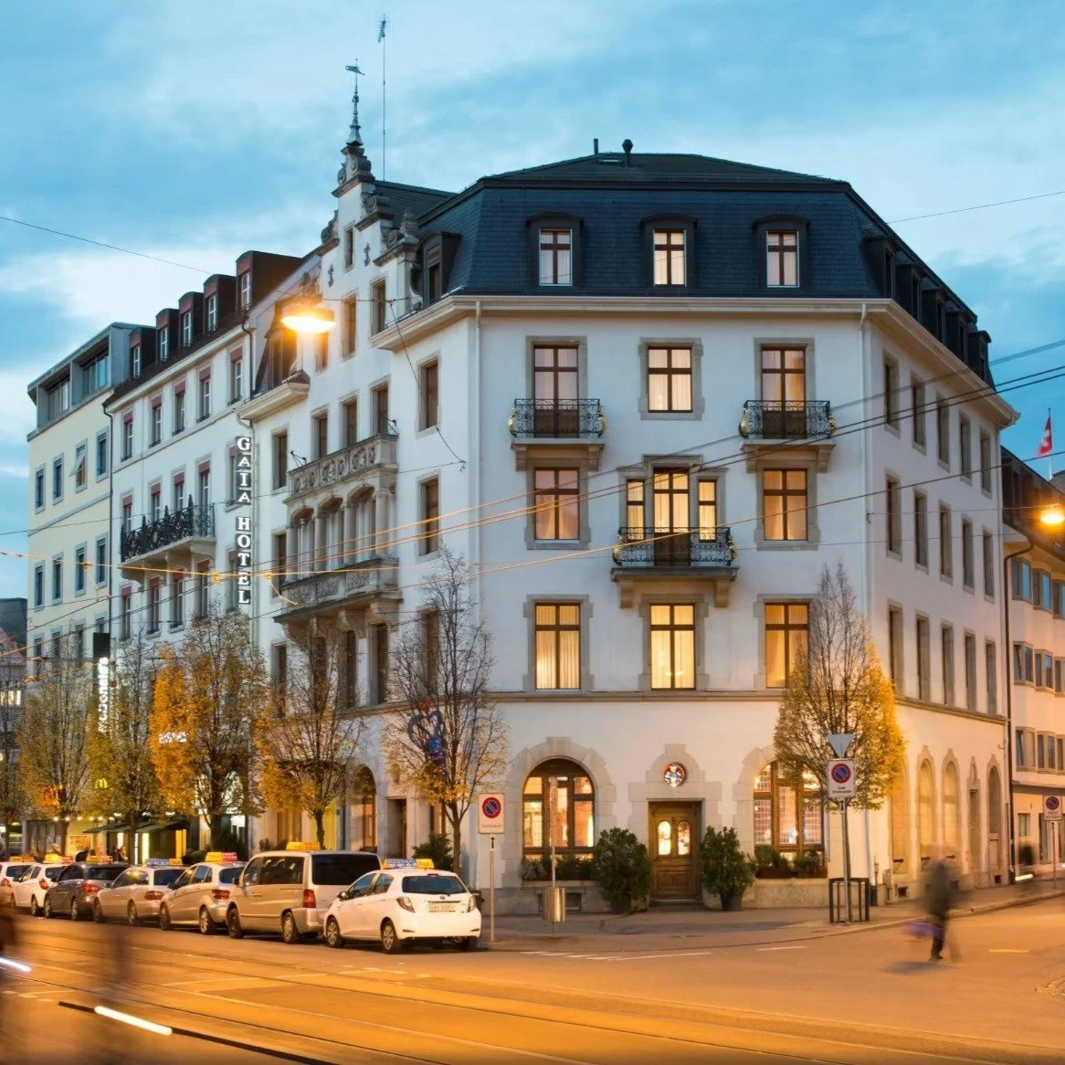 Ein mehrstöckiges Hotelgebäude namens GAIA HOTEL bei Sonnenuntergang, mit beleuchteten Fenstern, Straßenschildern, parkenden Autos und einigen Bäumen vor der Fassade.