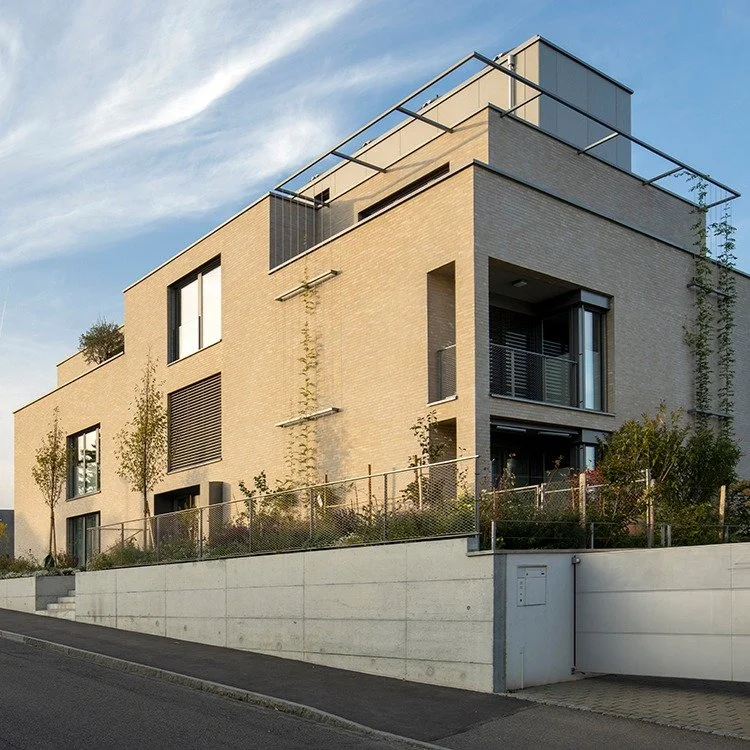 Modernes Mehrfamilienhaus mit Balkon und Gartenzaun, umgeben von Bäumen, bei Sonnenuntergang, in einer Wohnsiedlung in Deutschland.