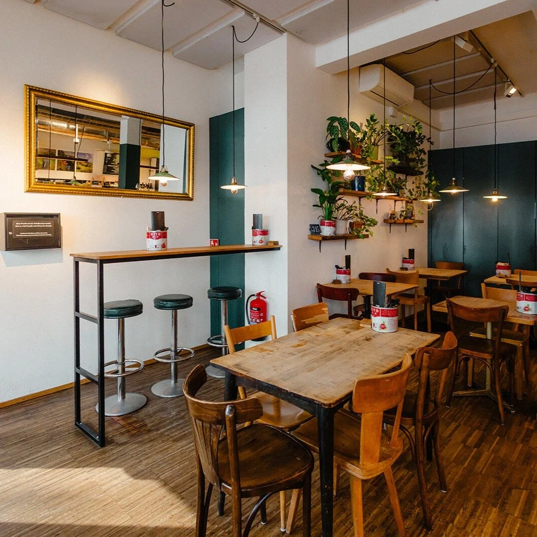 Innenraum eines Cafés mit Holztischen und Stühlen, grünen Wandakzenten, Pflanzen auf Regalen, Spiegel an der Wand, Deckenleuchten und Sicherheitsabständen auf den Tischen.