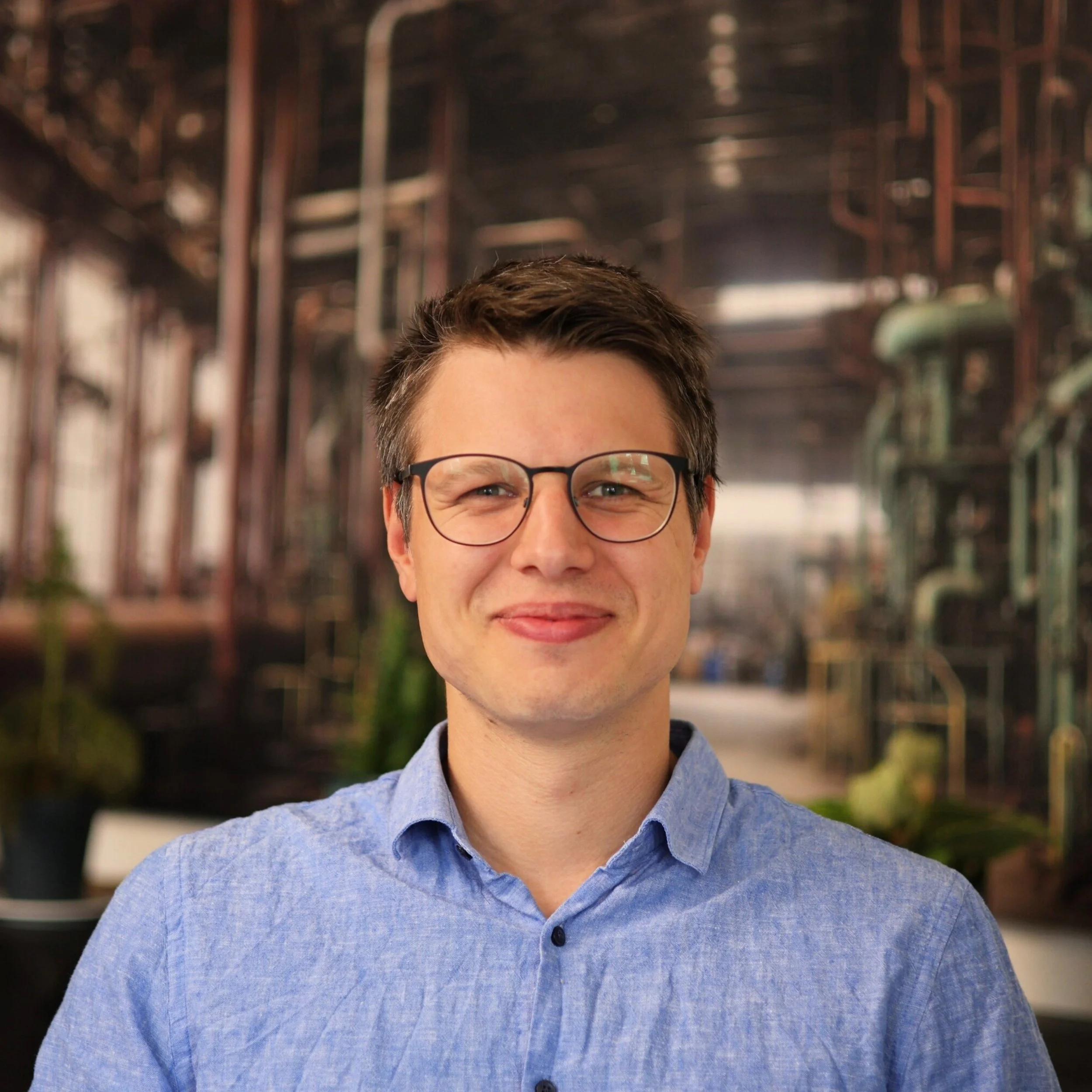 Ein Mann mit Brille und blauer Hemd, der vor einem Hintergrund mit mehreren Gärtanks und Pflanzen steht.