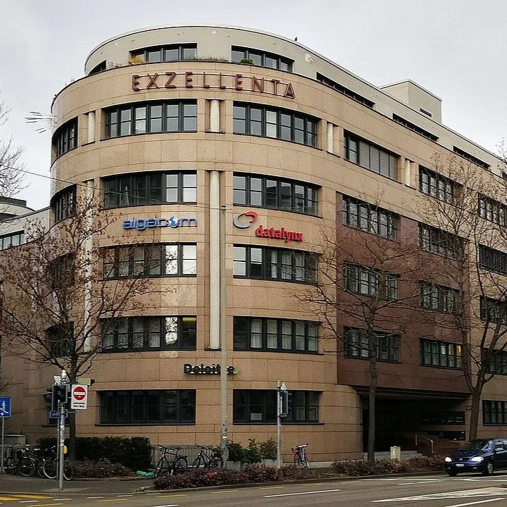 Mehrstöckiges Gebäude mit Firmenlogos und einem Hinweis 'Exzellenz' auf der Fassade, umgeben von Bäumen und Fahrradständern