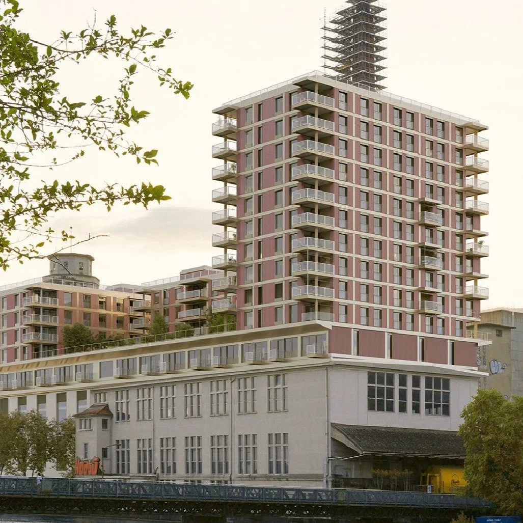 Mehrstöckiges modernes Wohngebäude mit zahlreichen Balkonen, im Hintergrund ein Fabrikgebäude und Bäume, in der Nähe eines Flusses.