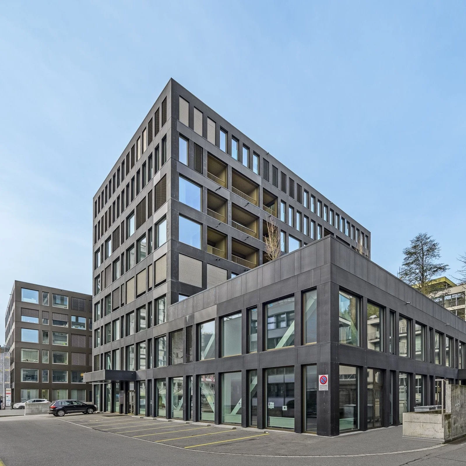Modernes schwarzes Bürogebäude mit vielen Fenstern und Balkonen gegen blauen Himmel, mit Parkplätzen und Bäumen im Vordergrund.