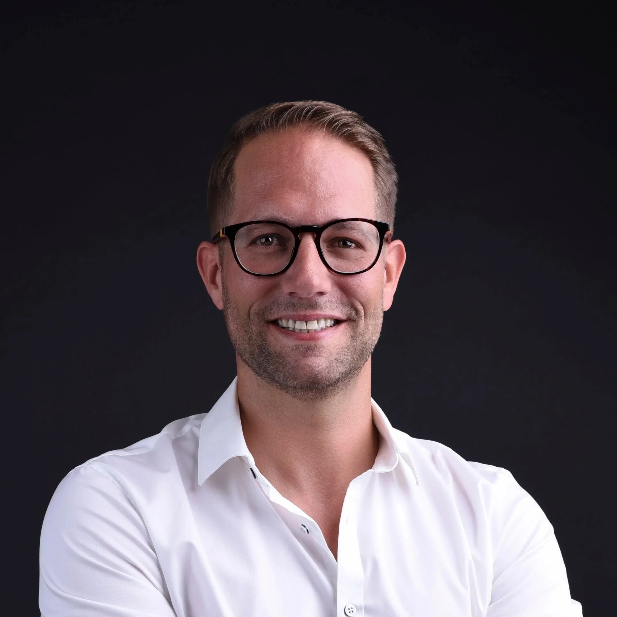 Nahaufnahme eines Mannes mit braunen Haaren, Brille und weihem Hemd vor schwarzem Hintergrund.