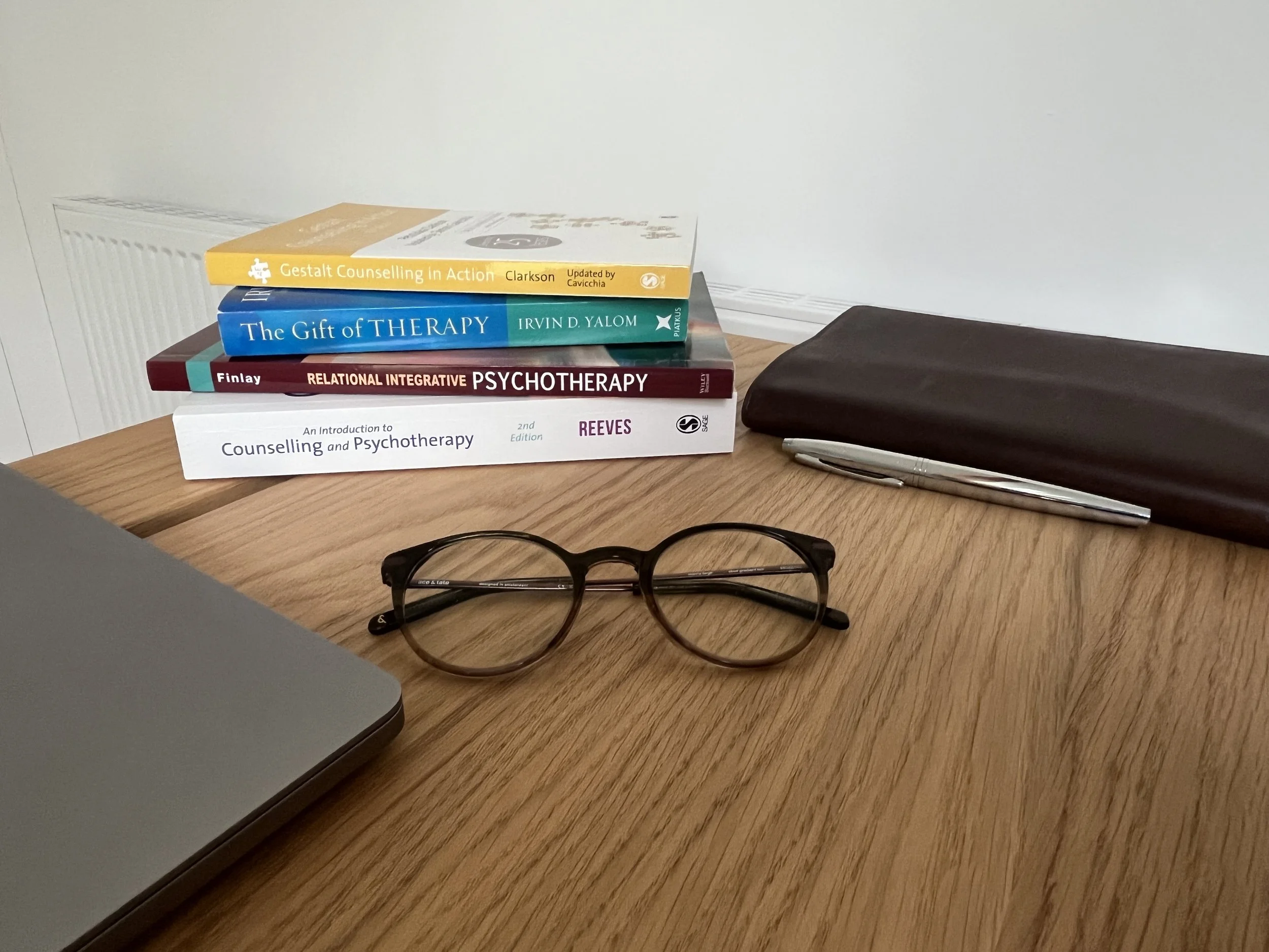 A wooden desk with four psychology books stacked on top, a closed laptop on the left, a pair of brown round eyeglasses in the center, a silver pen and a black leather notebook on the right.