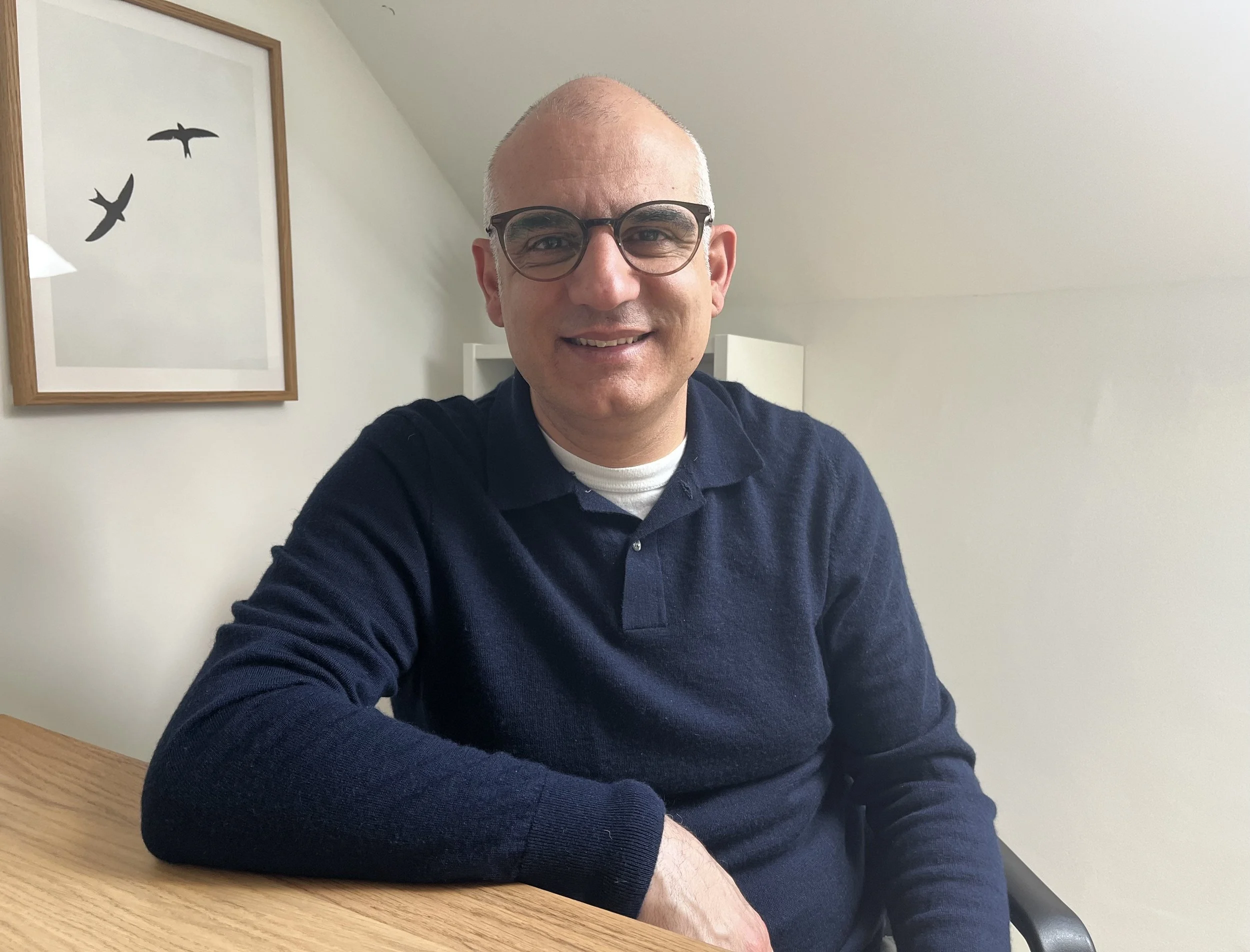 A smiling man with glasses and a bald head sitting at a table in a room with white walls and a framed picture of flying birds.