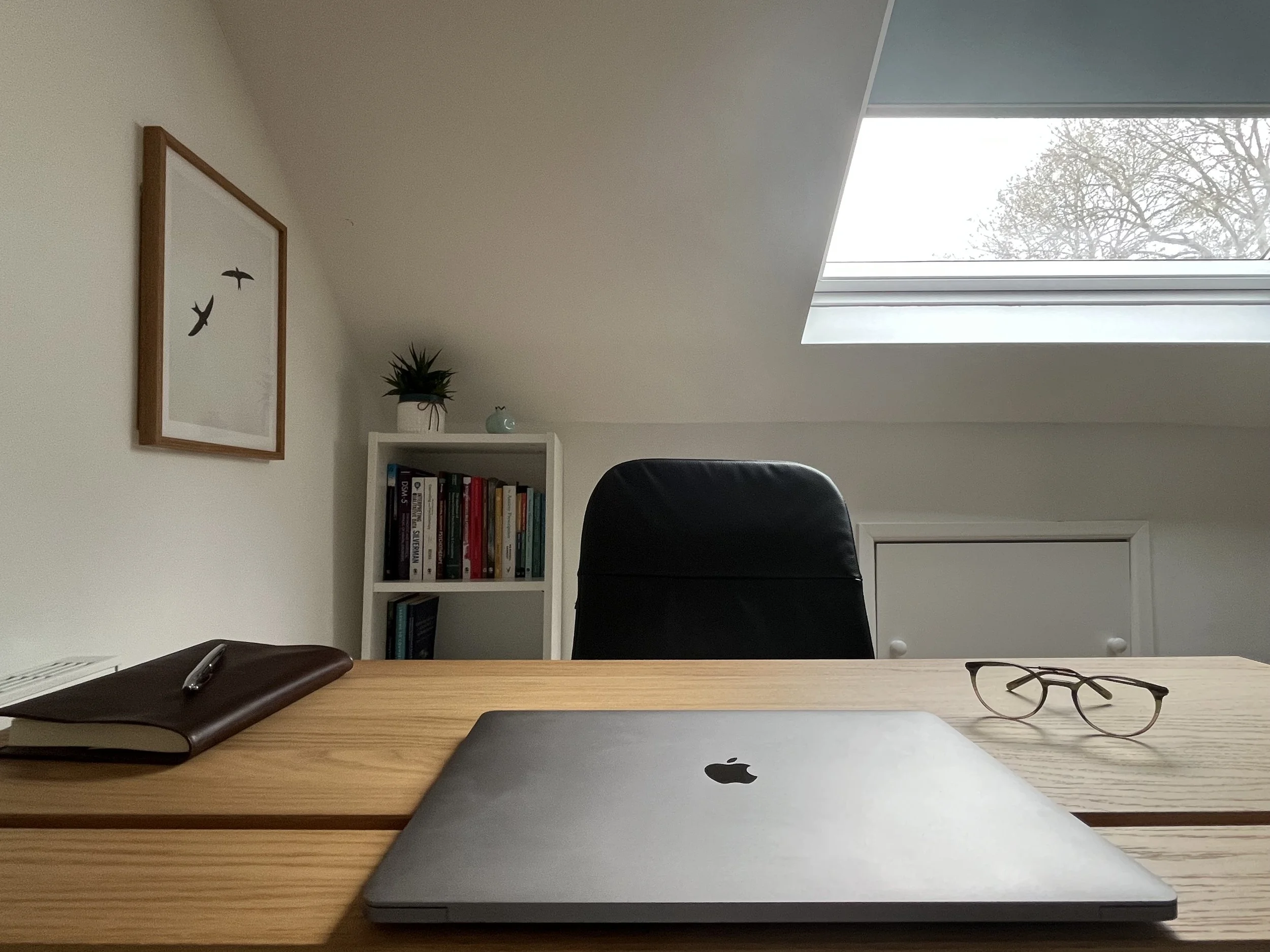 A desk in an attic office with a closed MacBook, glasses, a notebook with a pen, and a shelving unit with books and a potted plant, under a skylight window.