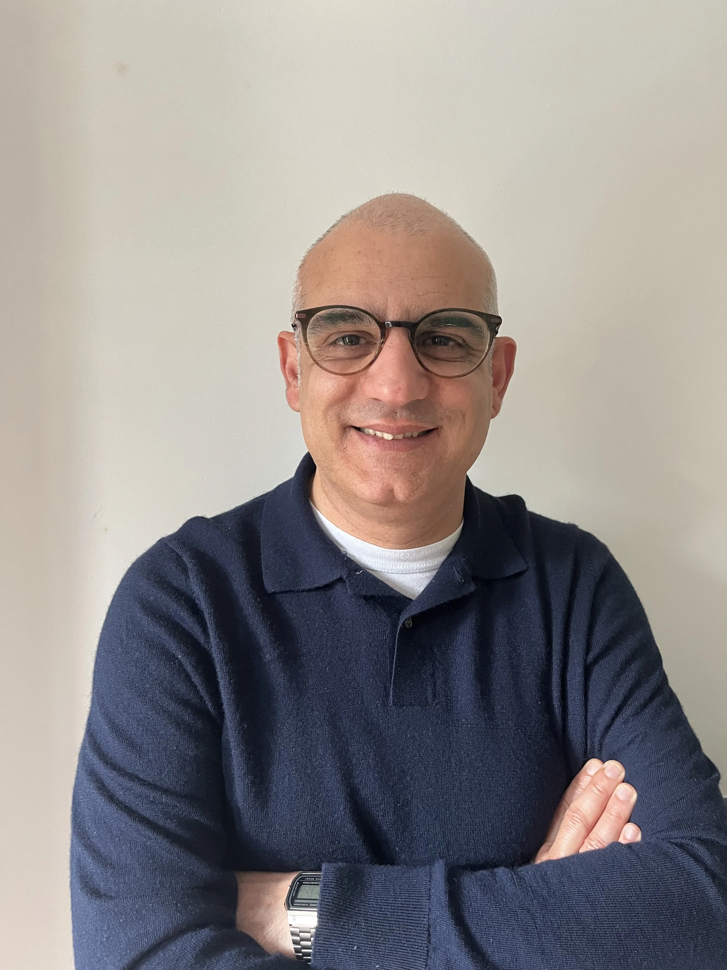 A man with glasses and a buzz cut hairstyle smiling with arms crossed, wearing a navy blue sweater and a watch, standing against a plain white wall.