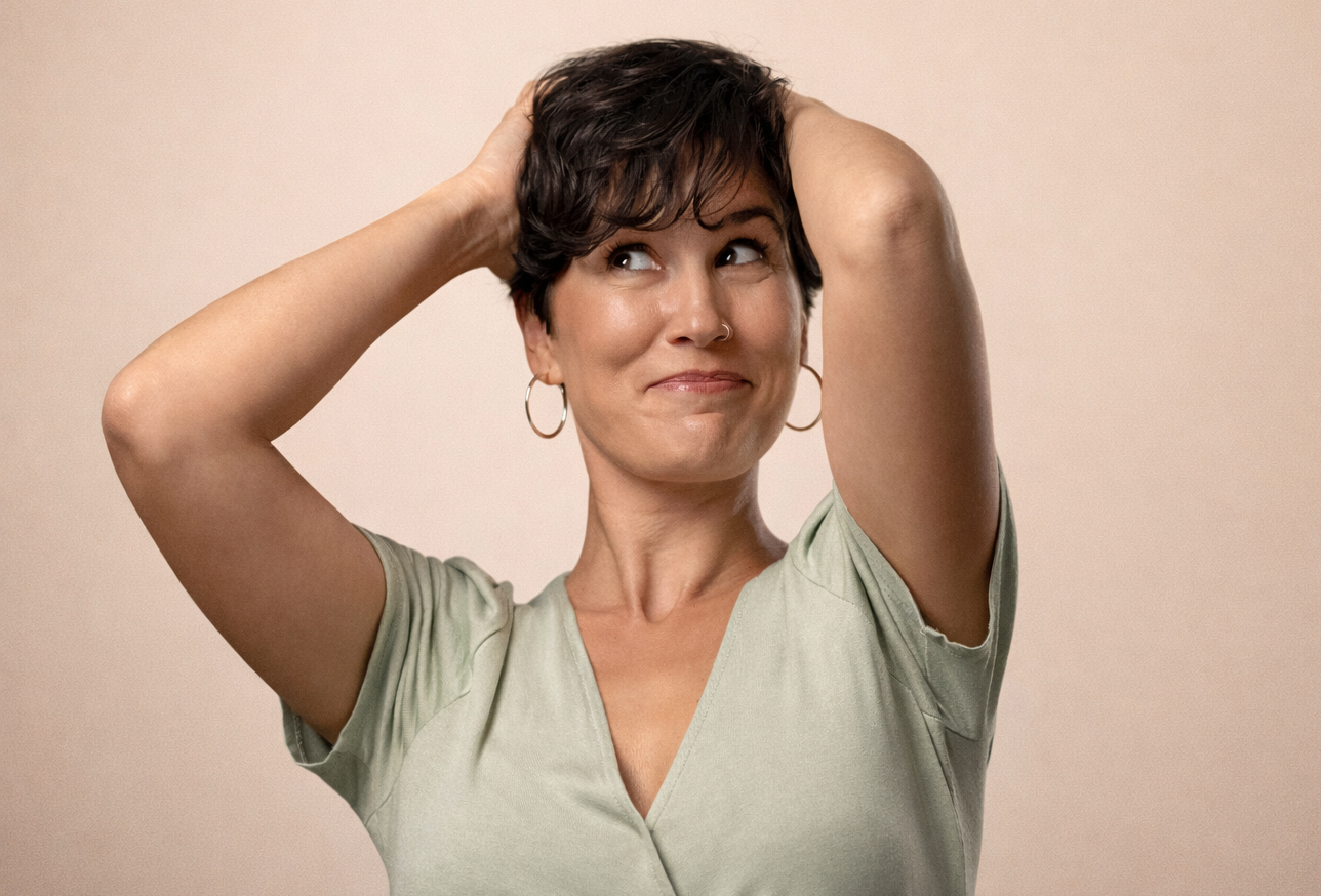 A woman with short dark hair and hoop earrings smiling with her hands on her head, wearing a light-colored V-neck shirt against a neutral background.
