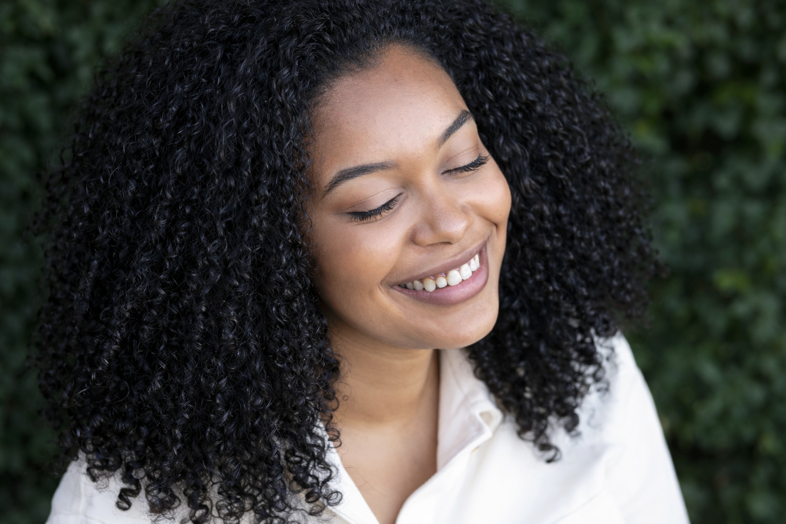 A woman with dark, curly hair smiling with her eyes closed outdoors.