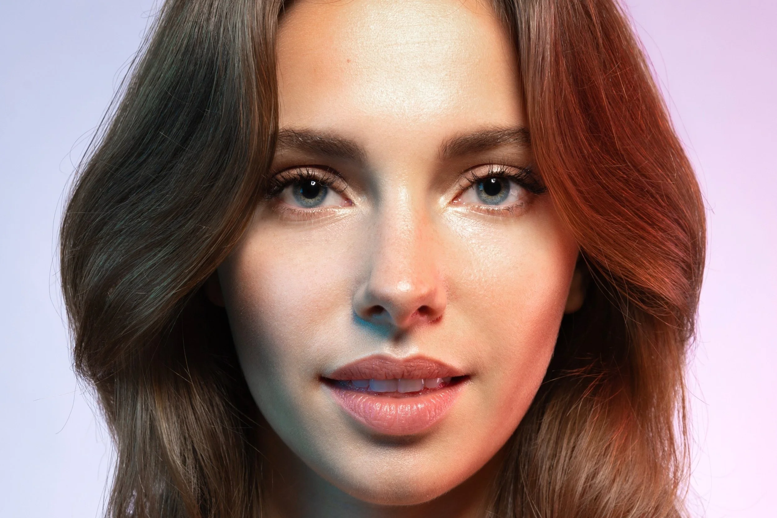 Close-up portrait of a young woman with long, wavy brown hair, blue eyes, and light makeup, looking at the camera with a slight smile.