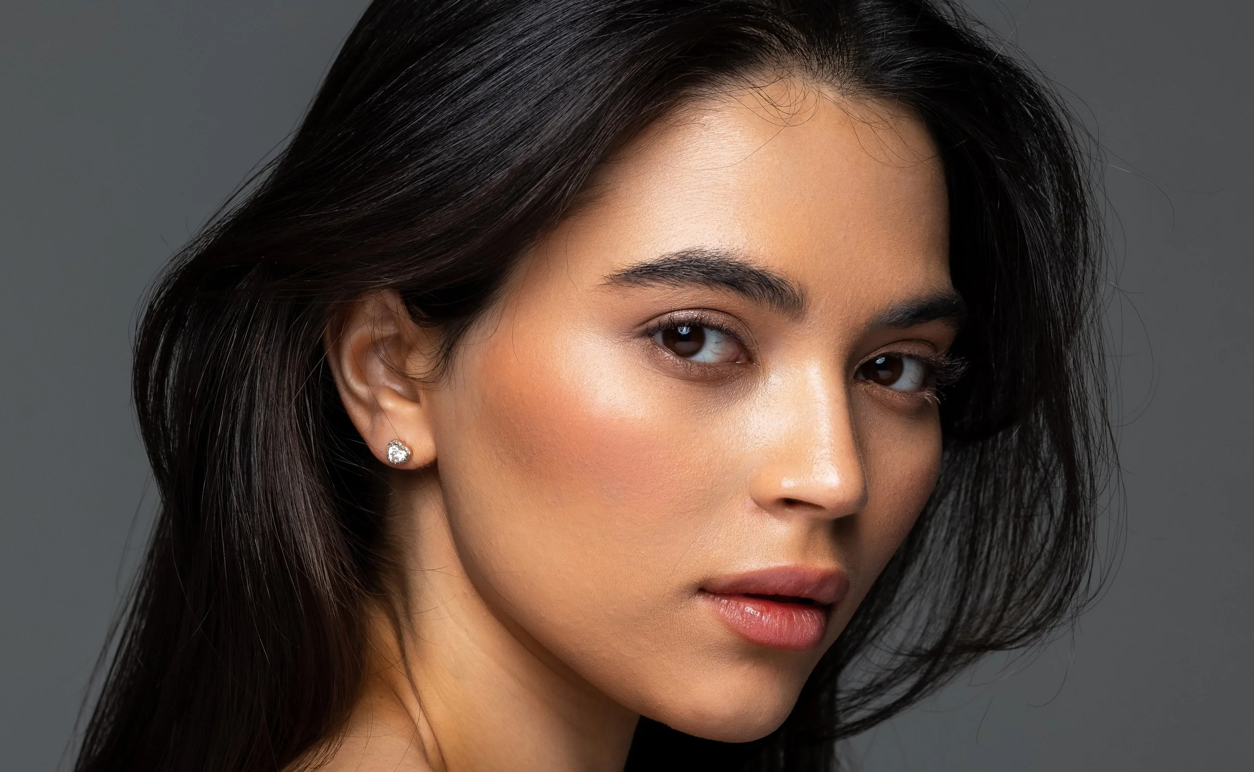 A close-up portrait of a young woman with fair skin, long dark hair, and wearing diamond stud earrings, against a gray background.