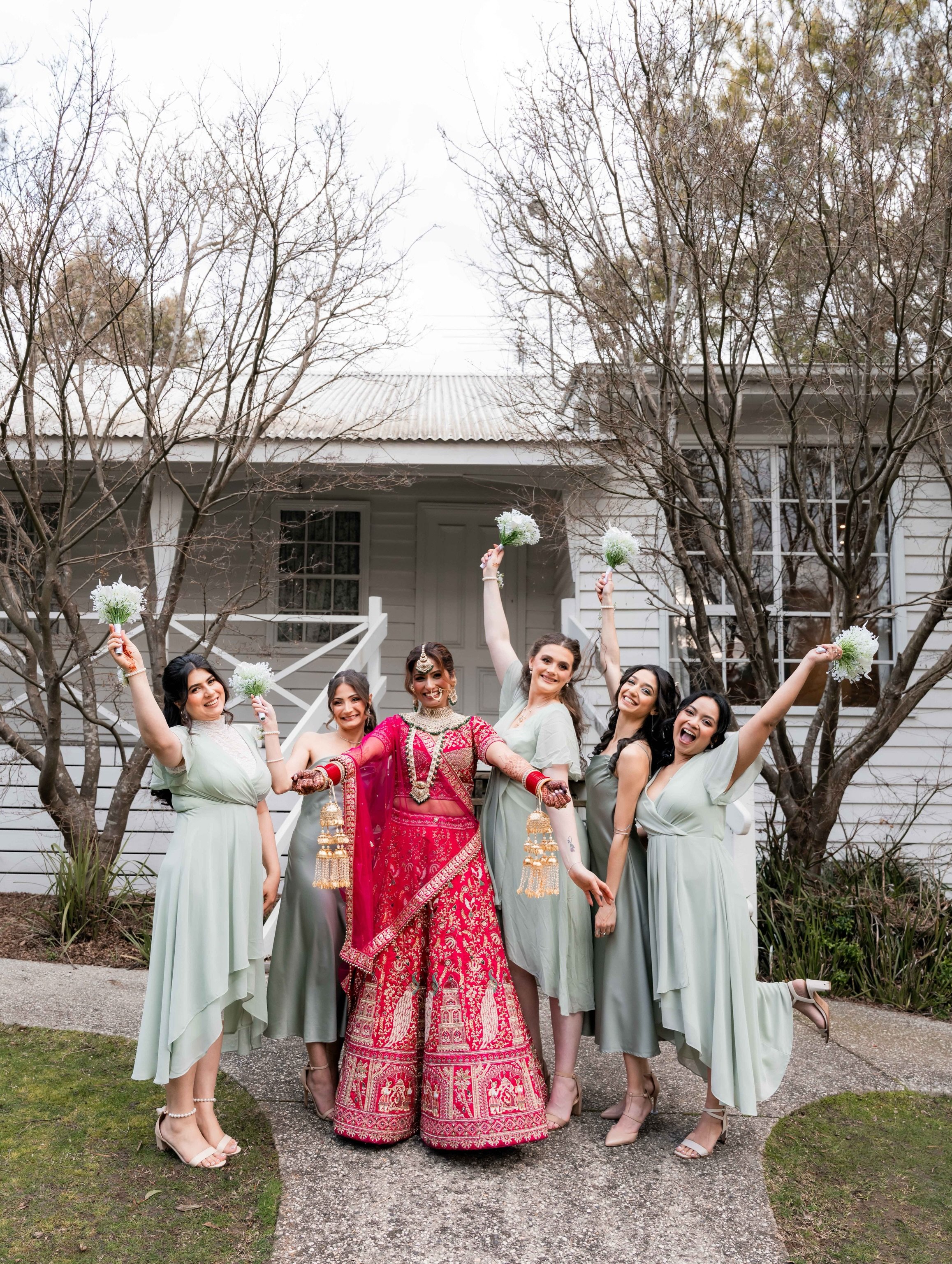 A group of women in formal attire, with the central figure wearing a traditional red and gold Indian dress, holding gold and pearl jewelry, and surrounded by women in pastel green dresses, celebrating outdoors in front of a house with leafless trees.