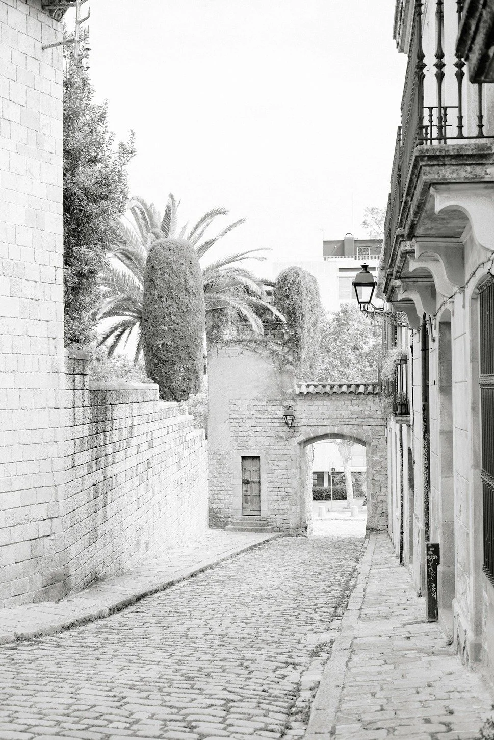 A narrow cobblestone street in an urban area, lined with buildings and trees, with an archway at the end, in black and white.