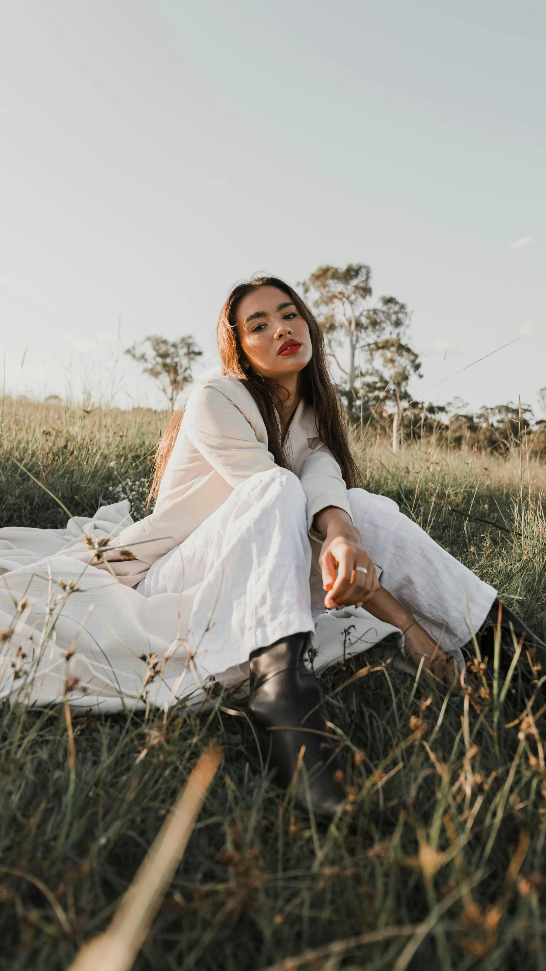 A woman with dark hair and red lipstick sitting on grass in a field, wearing a cream-colored jacket, white pants, and black boots, with trees and a clear sky in the background.