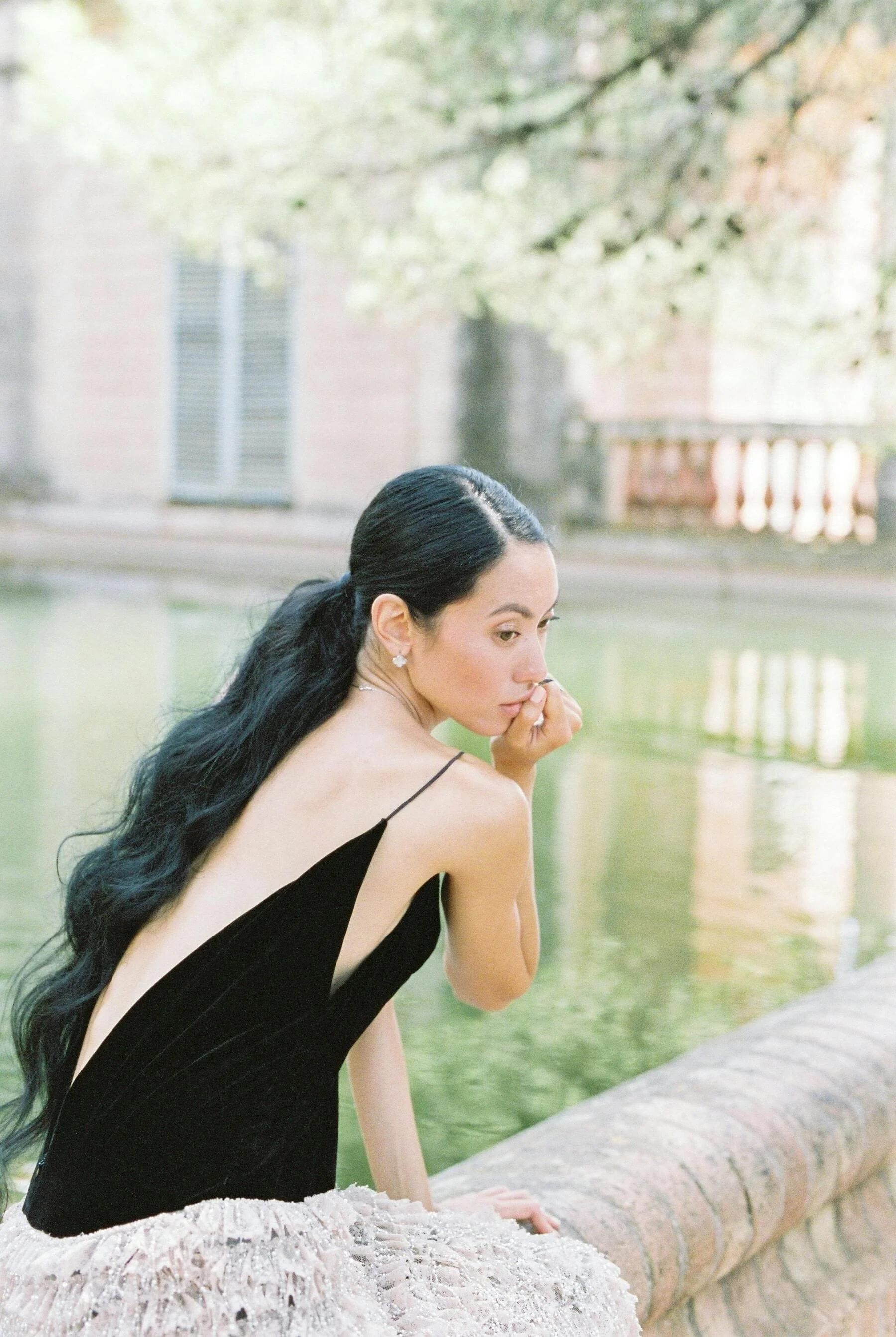 A woman with long black hair in a black dress, sitting on a stone wall by a river, with trees and buildings in the background, looking thoughtfully to the side.
