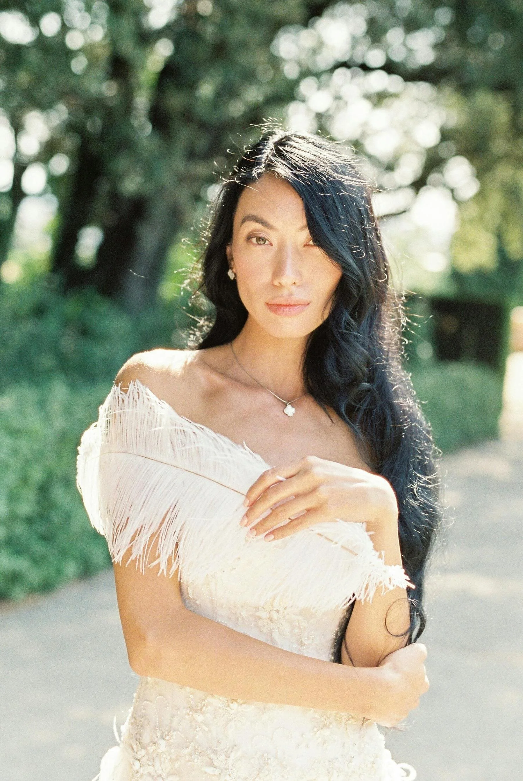 A woman with long black hair and light skin, wearing a white dress with fringe, standing outdoors on a sunny day with trees in the background.