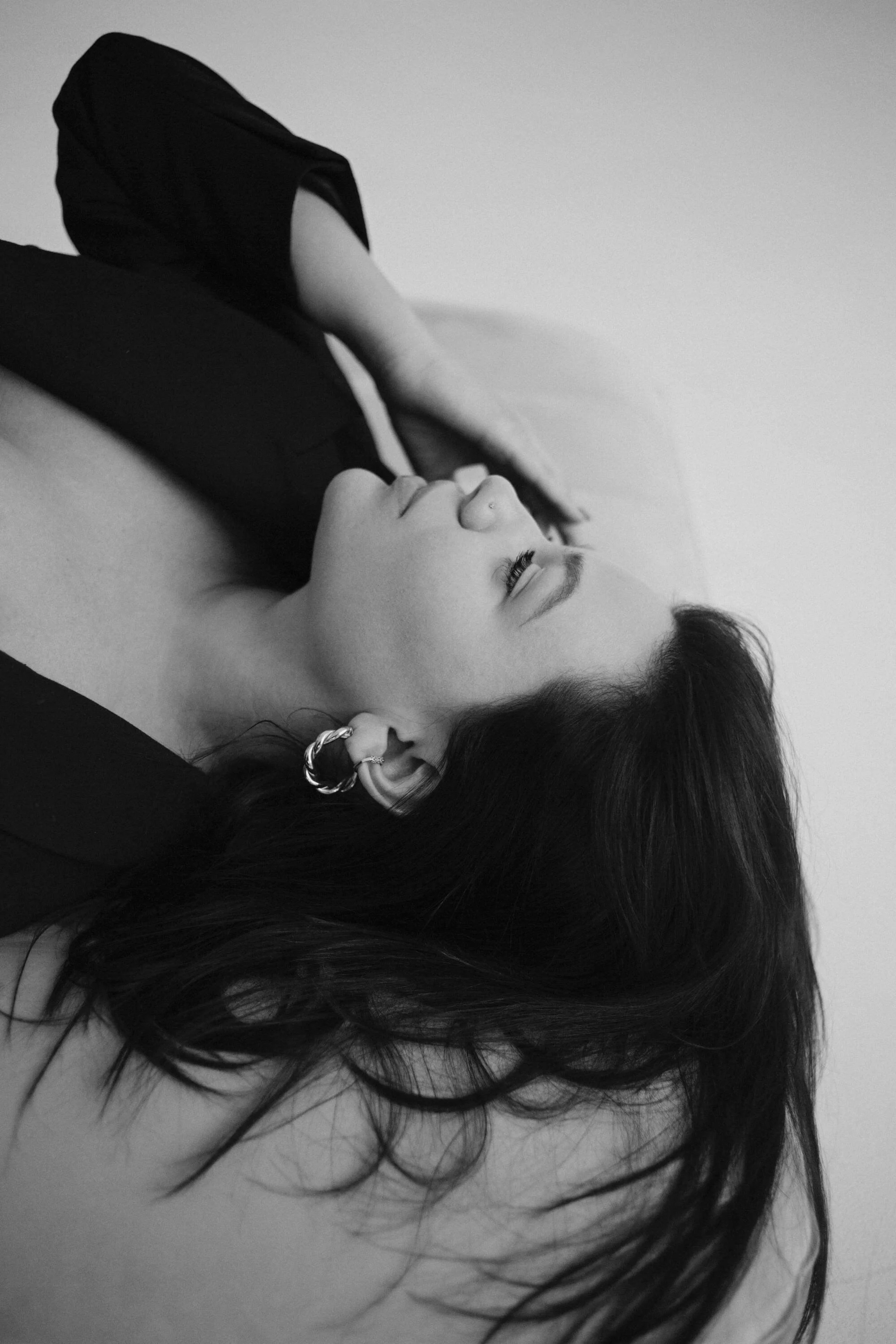 A woman with long dark hair wearing earrings, lying down on her back with her eyes closed, in a black and white photo.