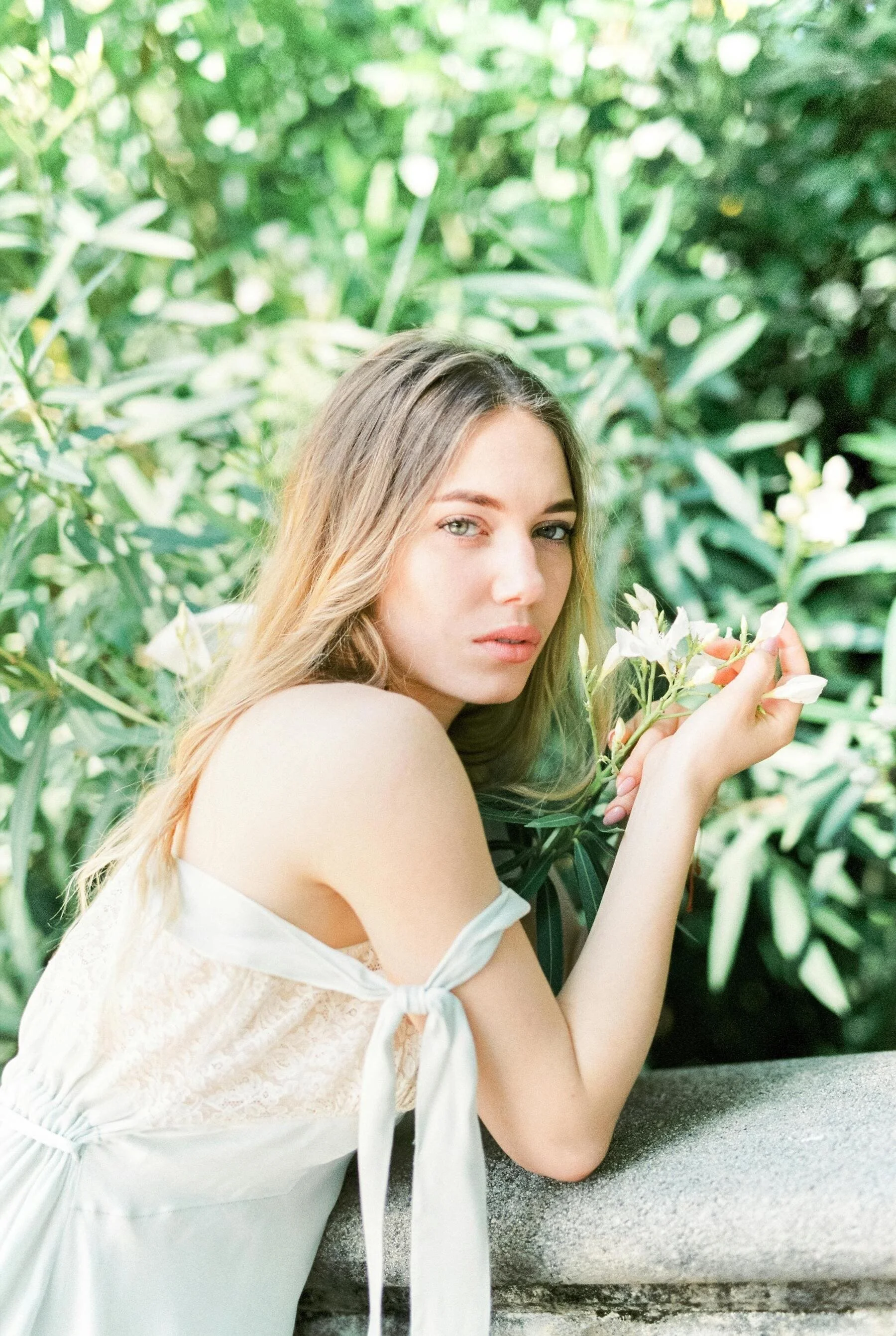 A young woman with long blonde hair holding white flowers in a garden with lush green foliage.