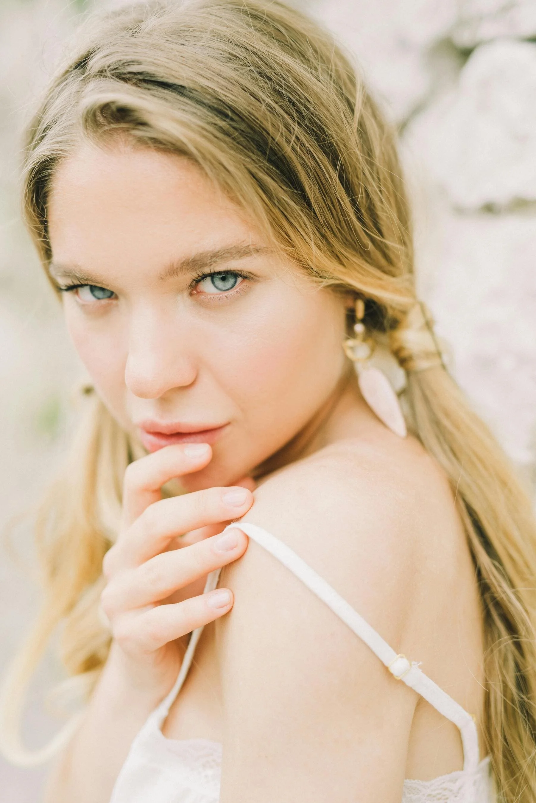 A young woman with blonde hair and blue eyes, wearing a white top, looking into the camera with one finger near her lips, outdoors with a blurred white wall background.