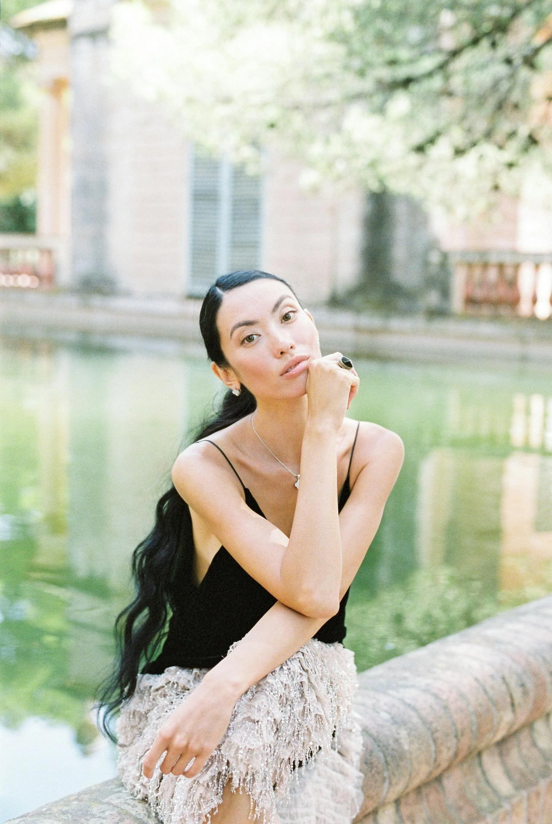 A woman with long dark hair, wearing a black spaghetti strap top and a beige lace skirt, posing by a stone ledge near a body of water, with greenery and trees in the background.