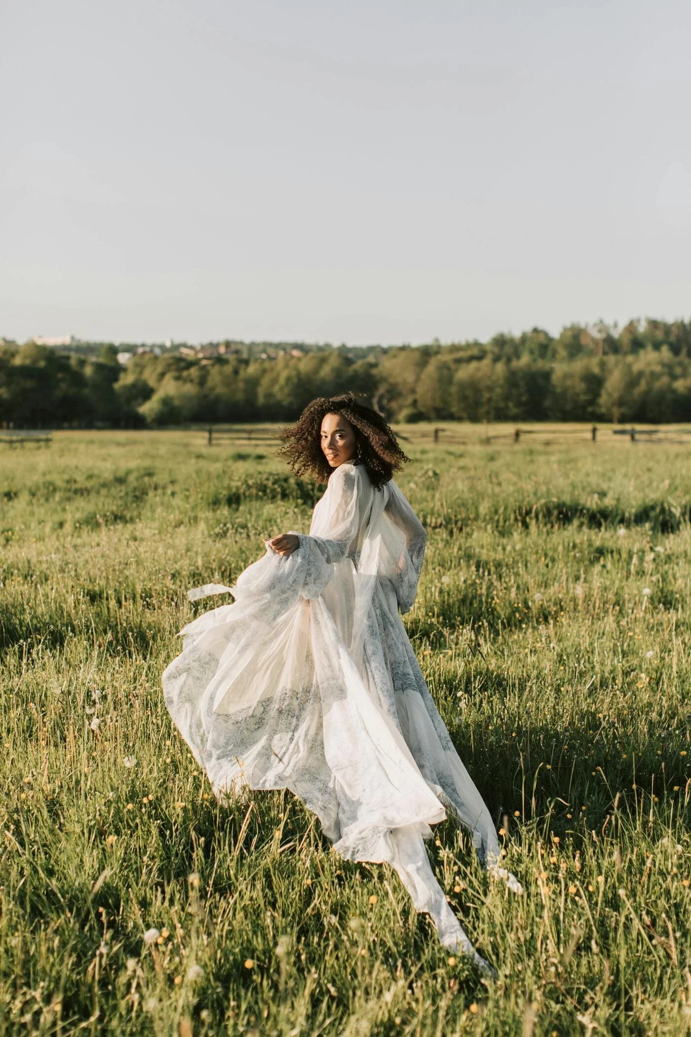 A woman in a flowing white dress standing in a grassy field during daylight.