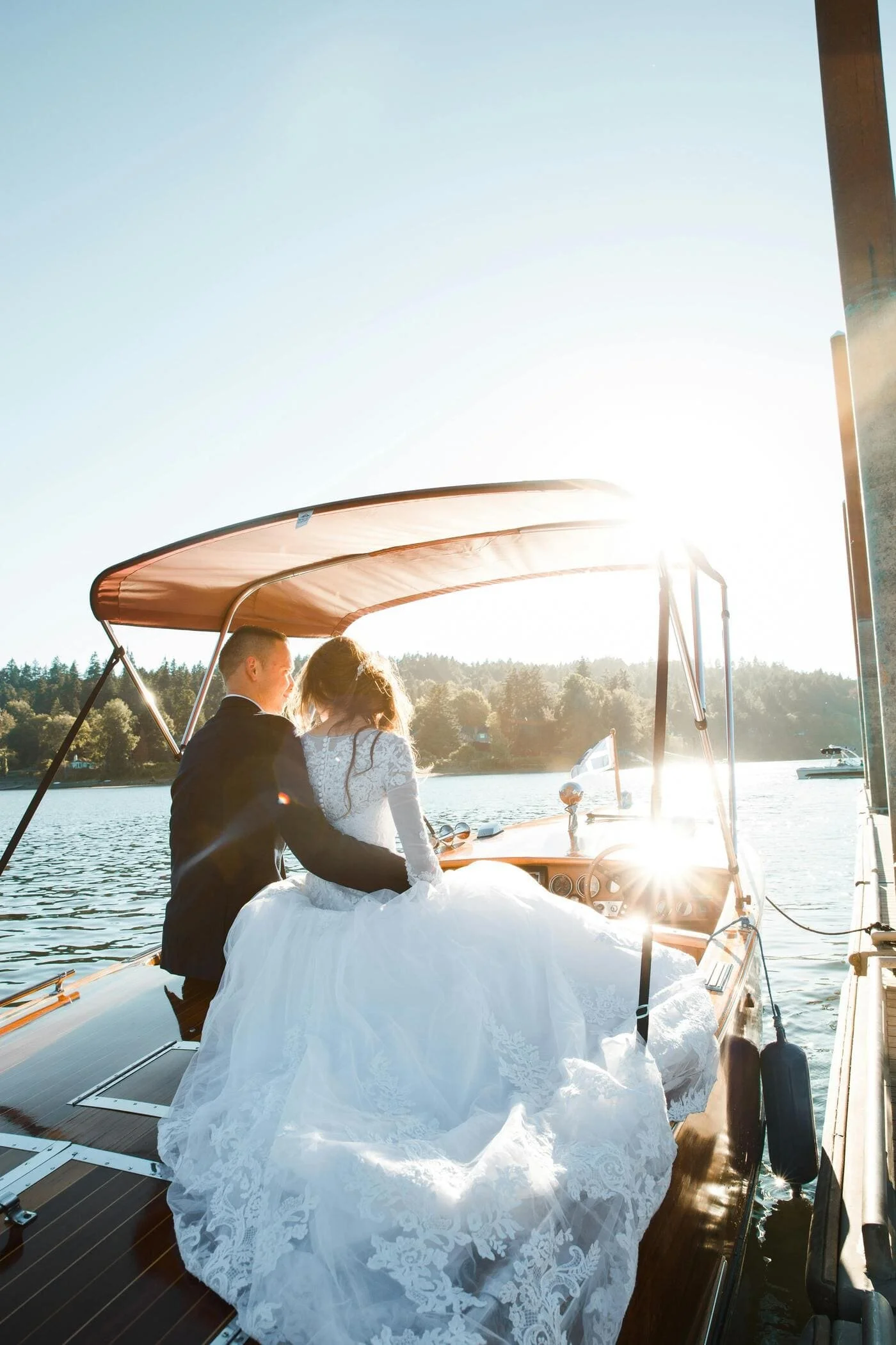 A bride and groom in wedding attire sitting together on a boat on a lake during sunset.