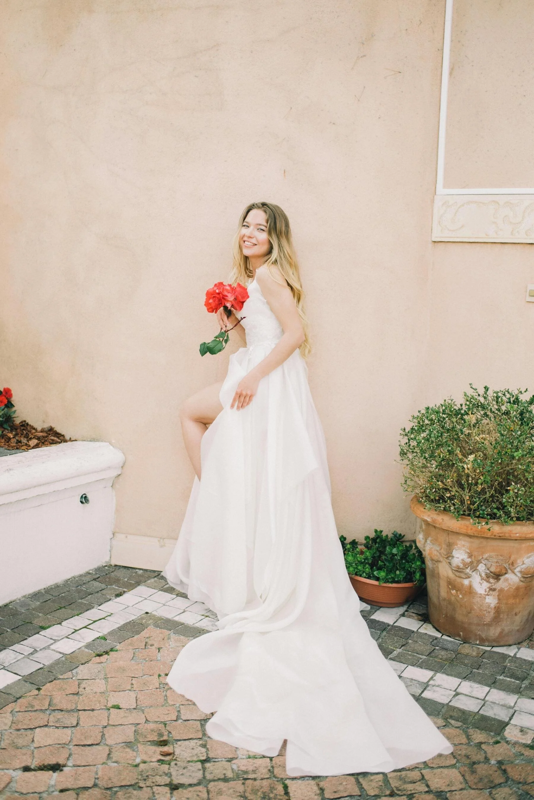 A woman in a wedding dress holding red flowers, standing outside on a cobblestone patio near potted plants, smiling at the camera.