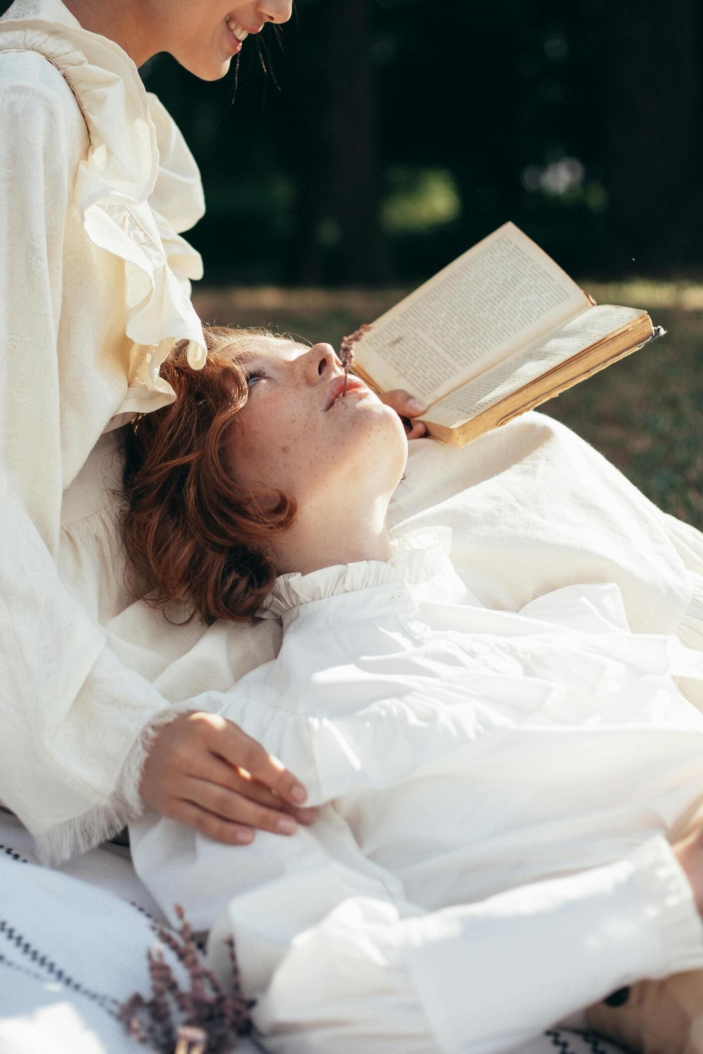 Two women relaxing outdoors: one lying on her back reading a book, the other leaning over her, smiling.