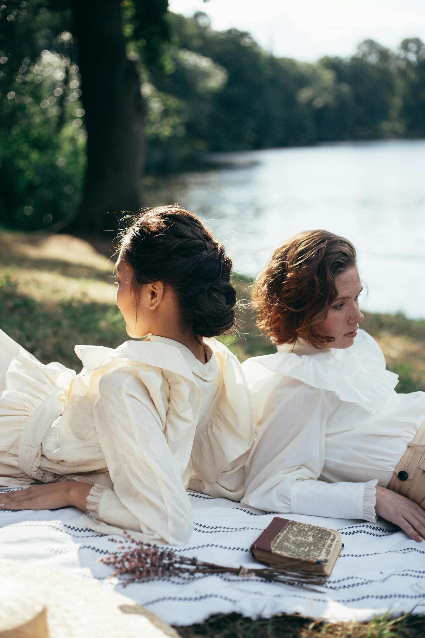 Two women with vintage clothing lying back to back on a picnic blanket by a river, with trees in the background, one reading a book.