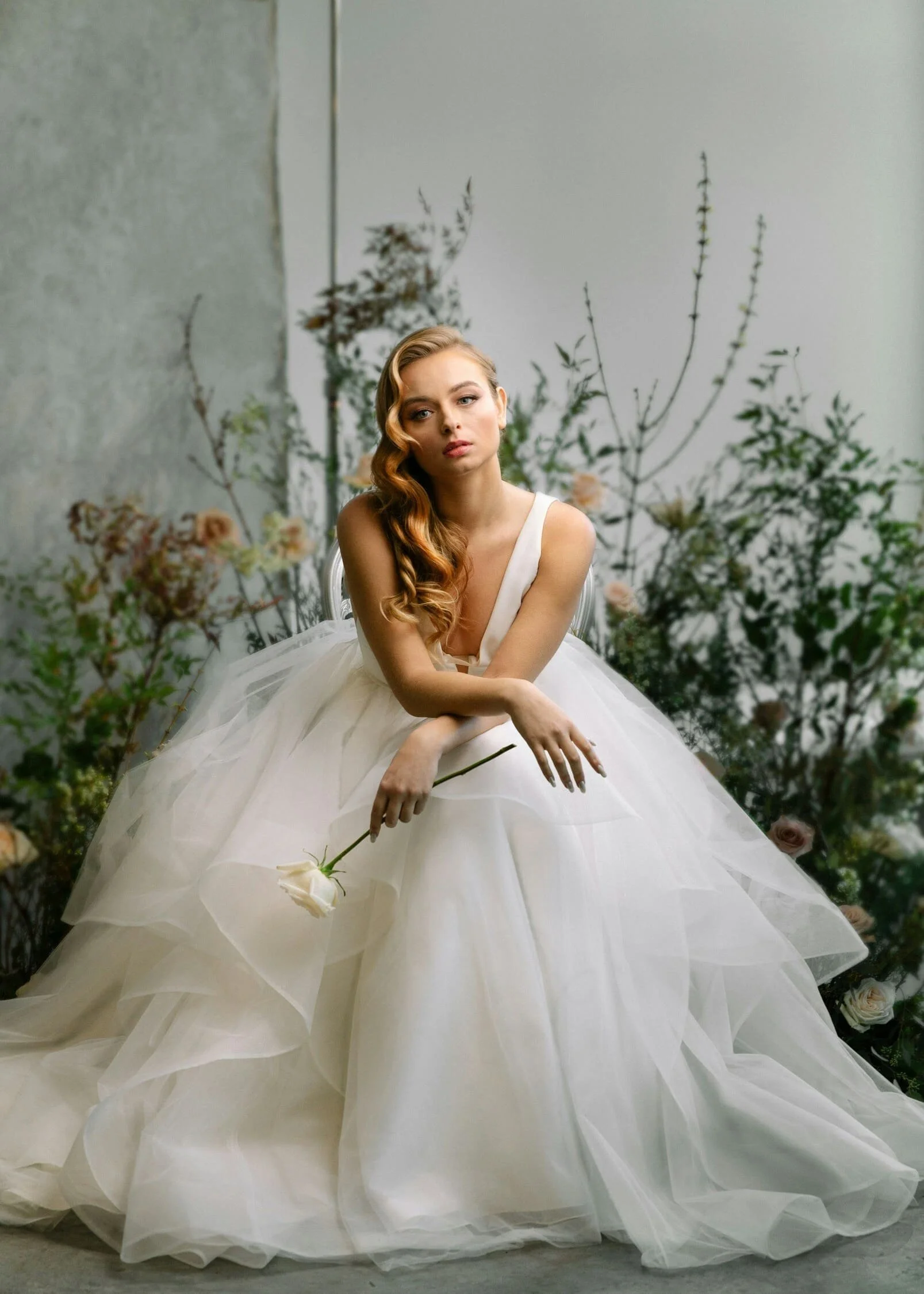 A woman in a white wedding dress sitting amidst a background of greenery and flowers, holding a single white rose.