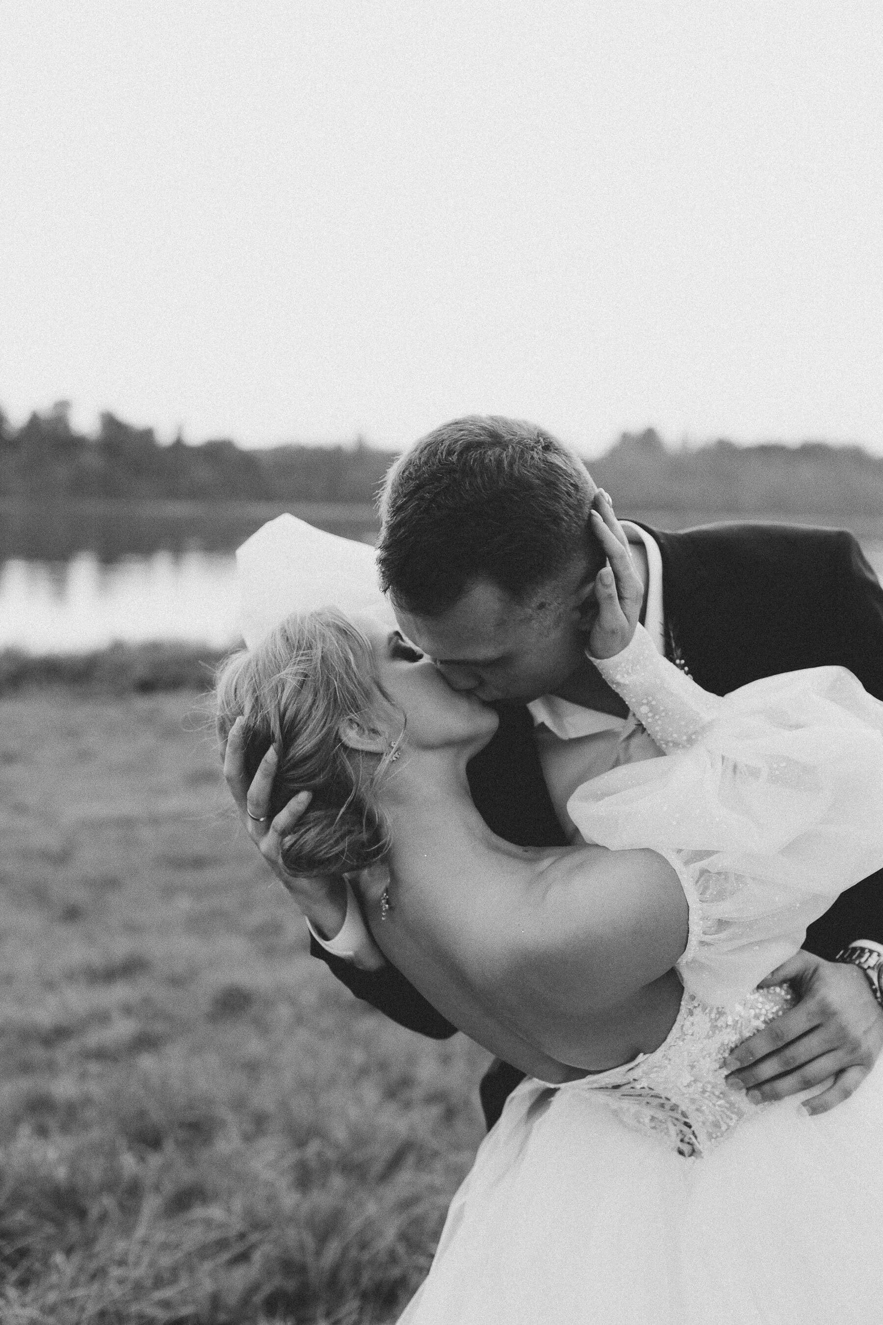 A black and white photo of a bride and groom sharing a kiss outdoors near a body of water, with the groom holding the bride and both embracing.