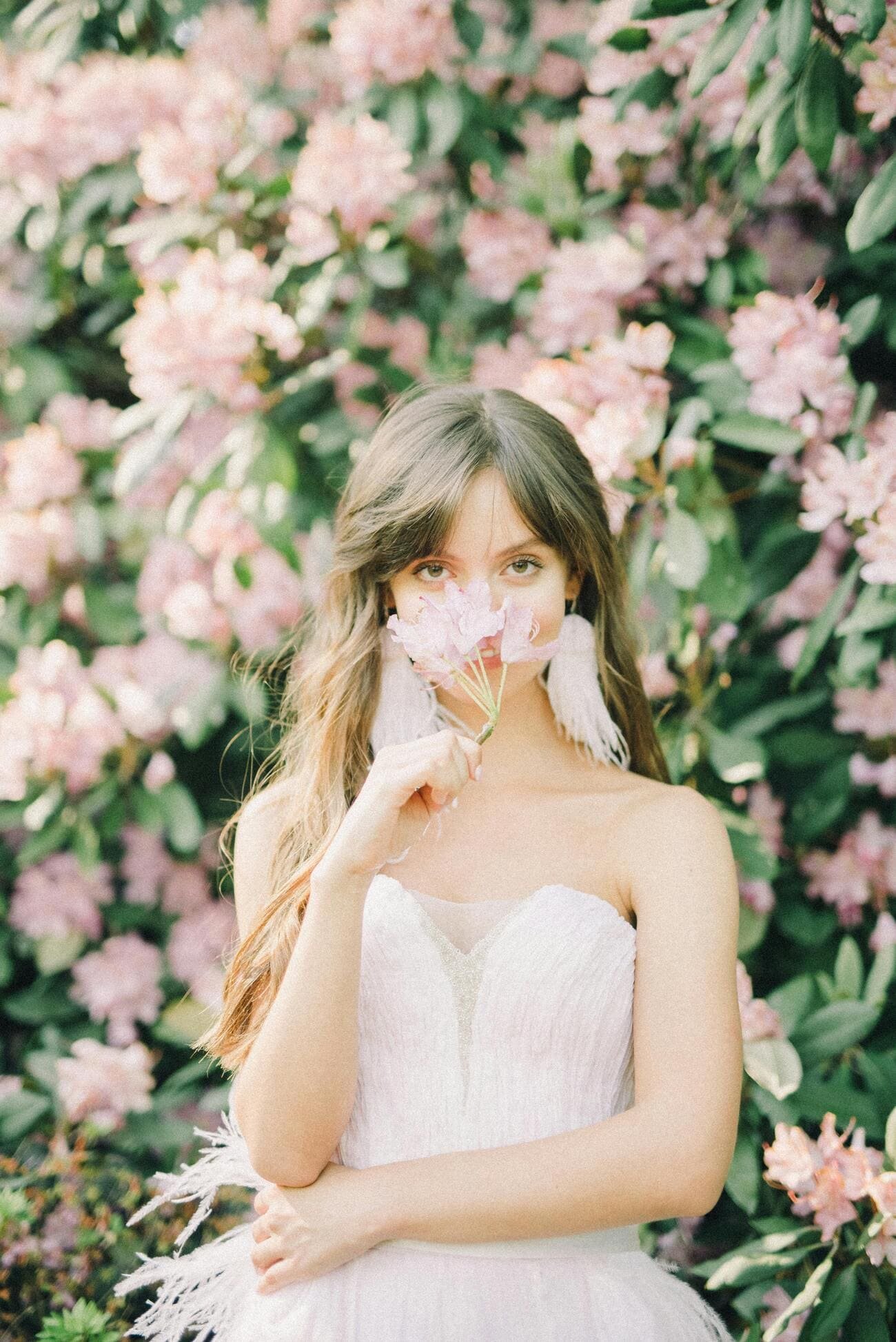 A young woman in a white dress holding pink flowers in front of her face, surrounded by pink and green flowers.