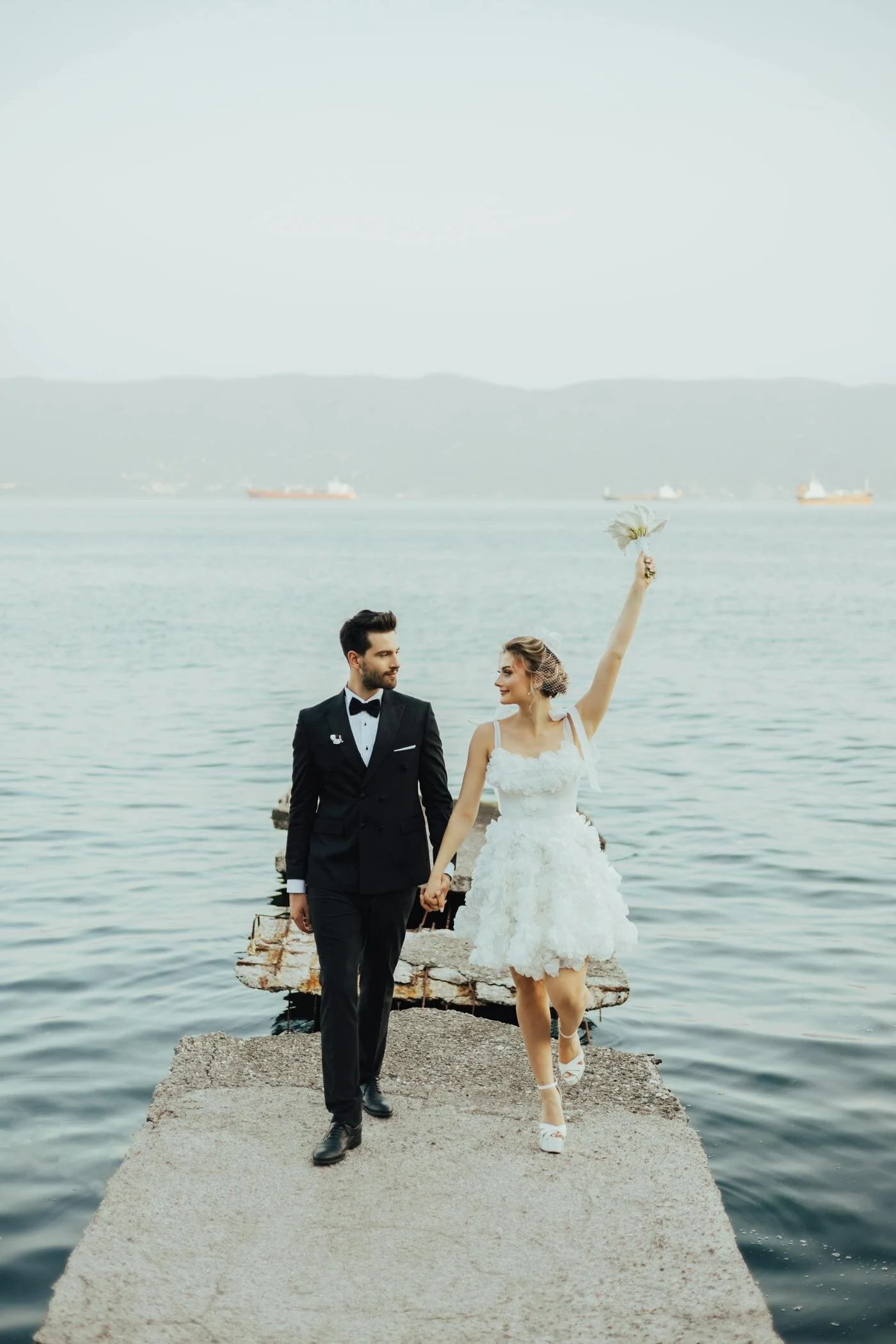 A newlywed couple walking on a concrete pier by the water, holding hands with the bride raising a bouquet in the air, against a backdrop of a large body of water and distant ships and hills.