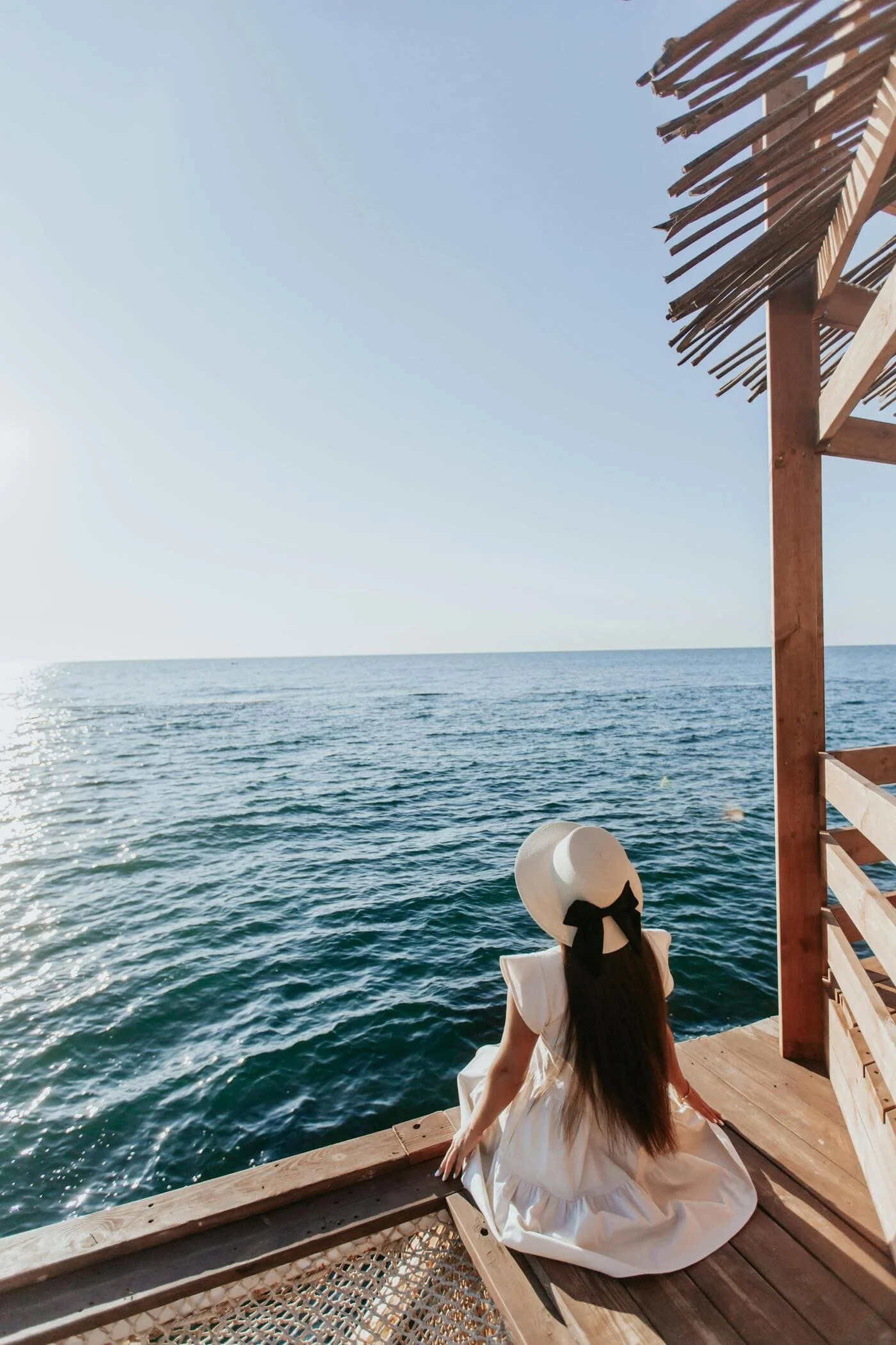 Woman in white dress and wide-brimmed hat sitting on wooden deck overlooking the ocean, with a thatched roof structure nearby.
