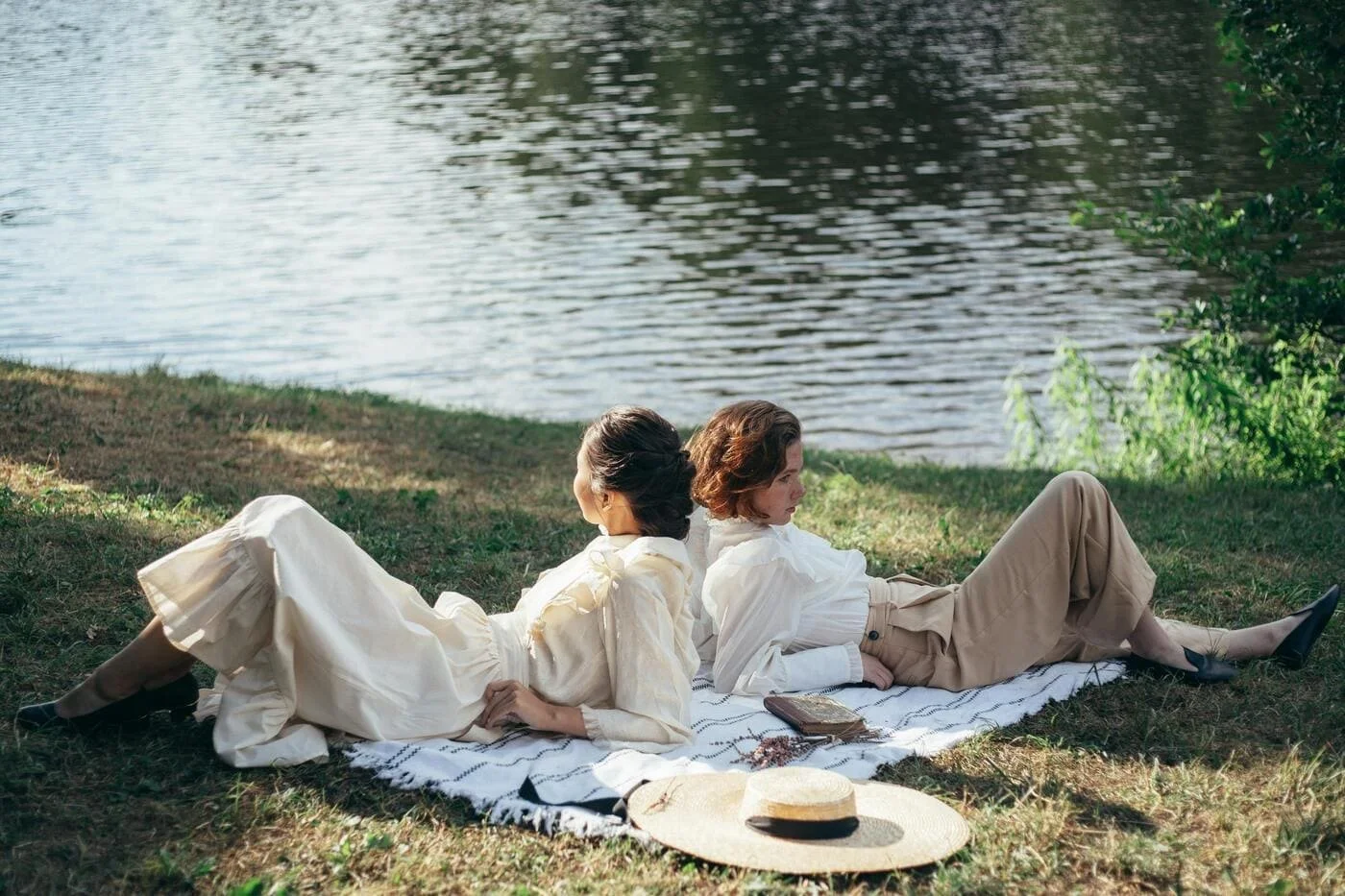 Two women in white tops and beige skirts or pants lying on a blanket near a body of water with trees, enjoying the outdoors on a sunny day.