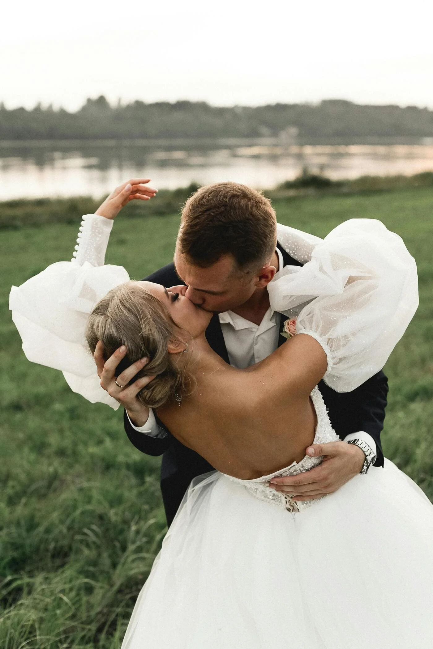 A bride and groom kissing outdoors near a lake during sunset, with the groom wearing a black suit and the bride in a strapless white wedding dress with puffed sleeves.