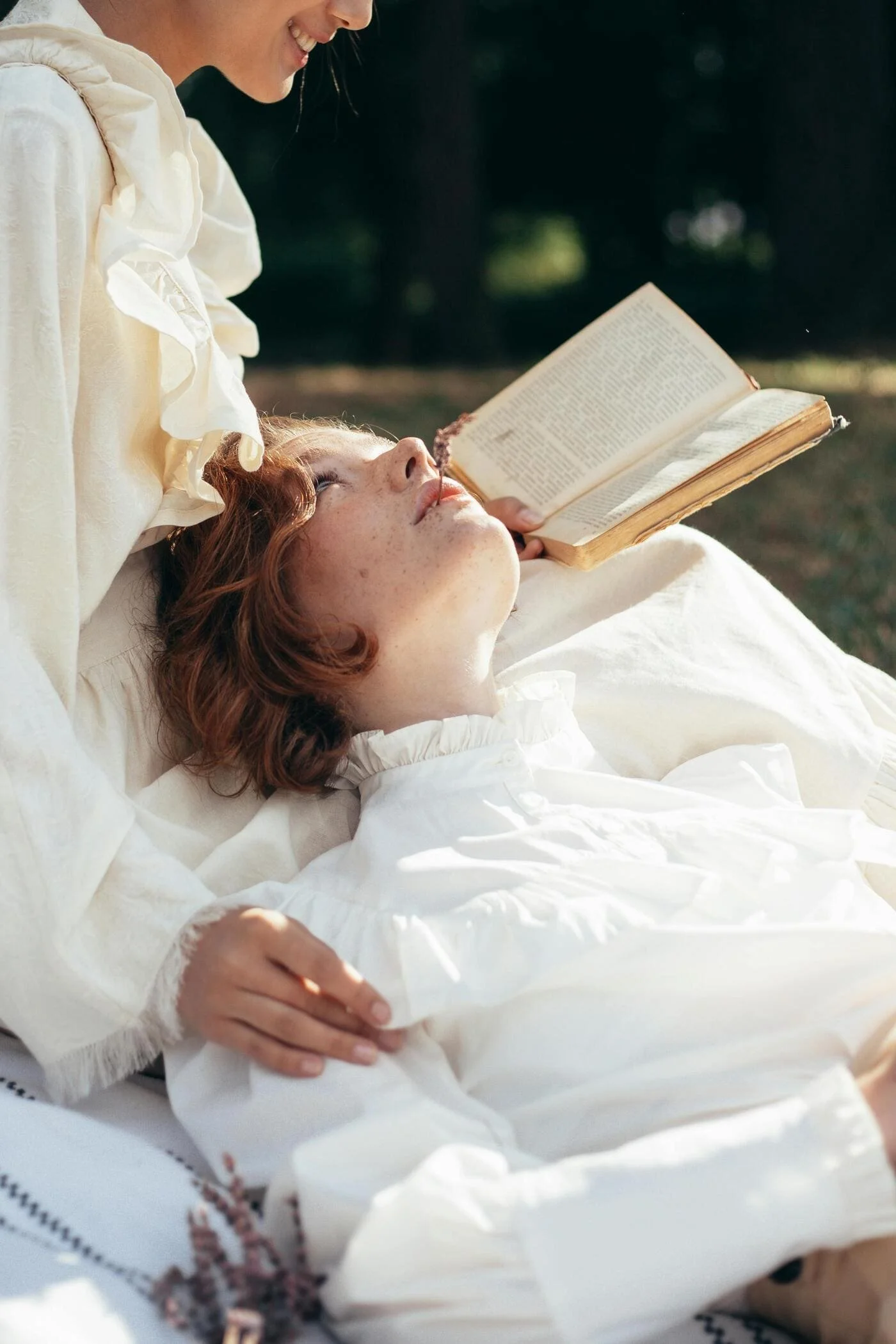 Two women, dressed in white, are outdoors with one lying on her back and the other leaning over her, reading a book.