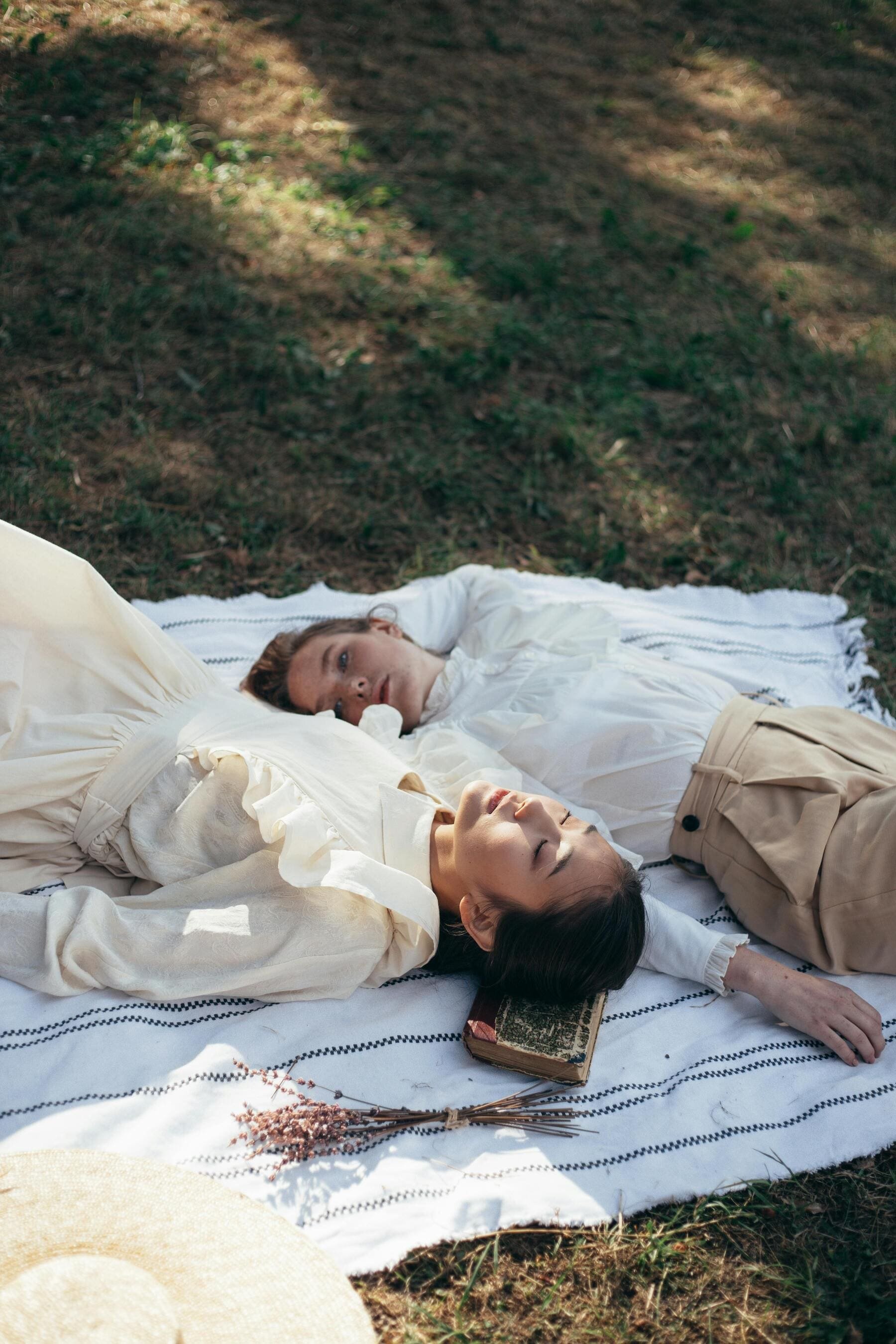 Two women are lying on a blanket spread on the ground outdoors, one with closed eyes and the other looking toward the camera. They are dressed in light-colored vintage-style clothing, surrounded by natural scenery, with a small bouquet of dried flowers nearby.
