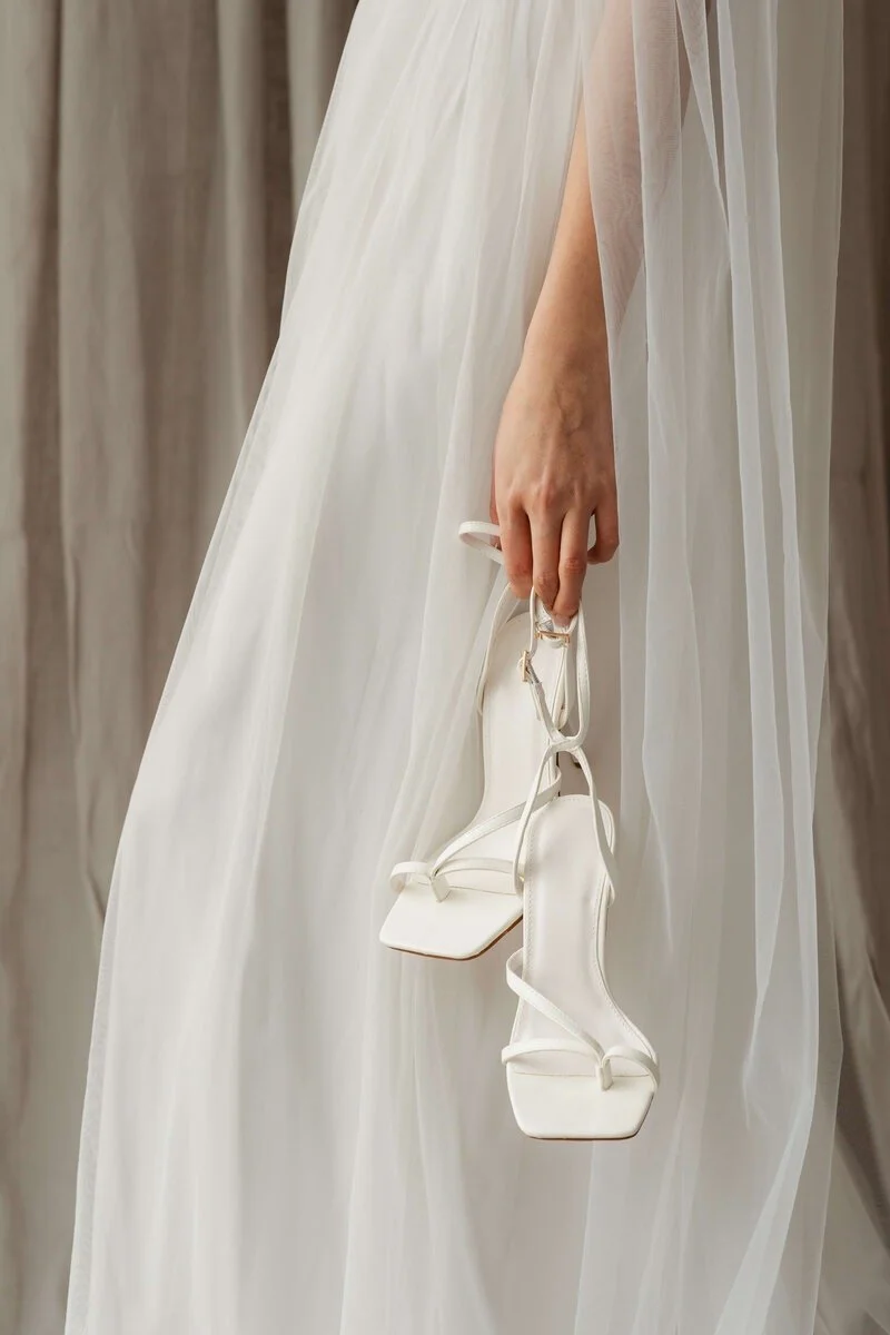 A person in a white dress holding a pair of white high-heeled sandals.