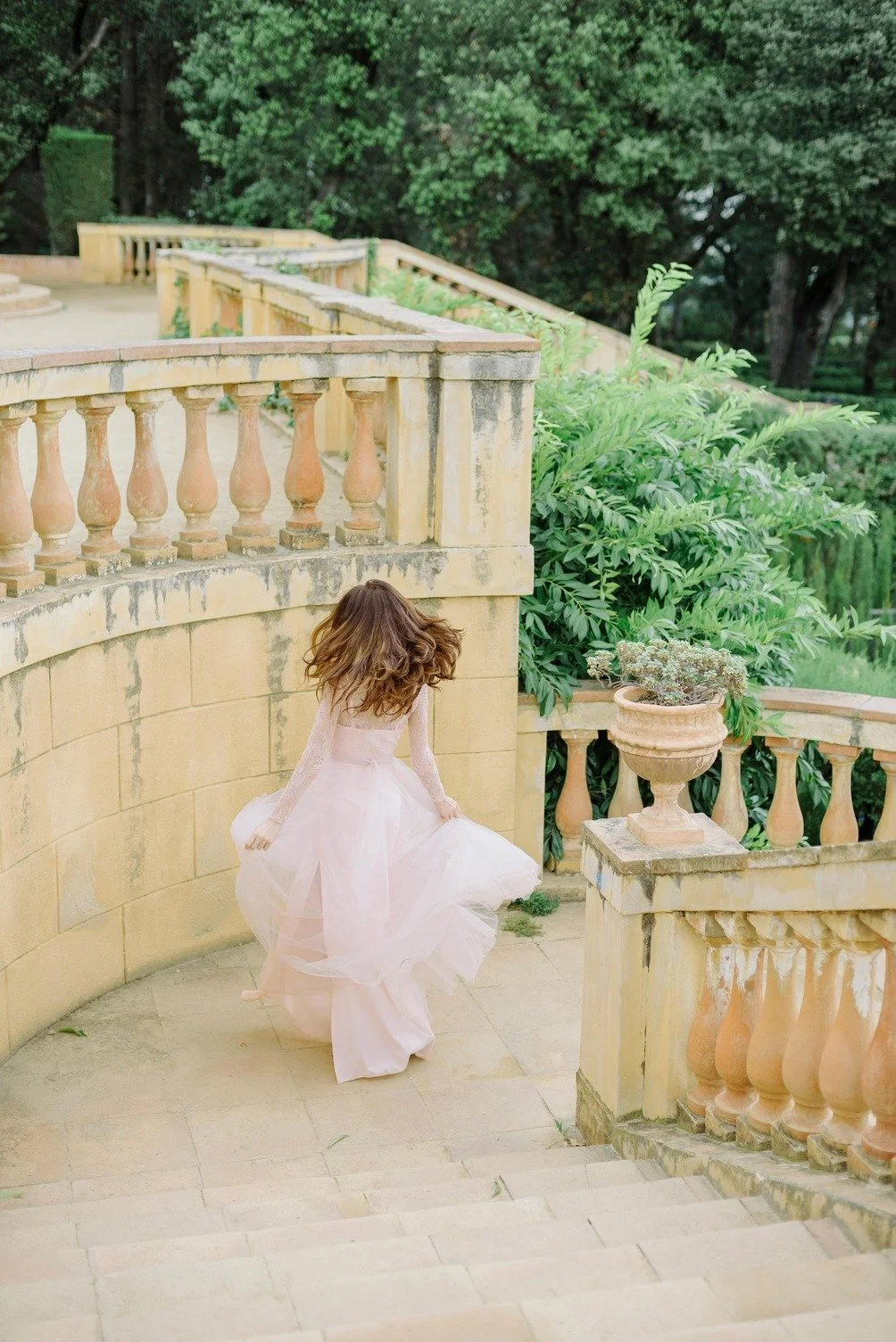 A woman in a flowing pink dress walking down stone stairs with a curved stone wall and lush green trees in the background.