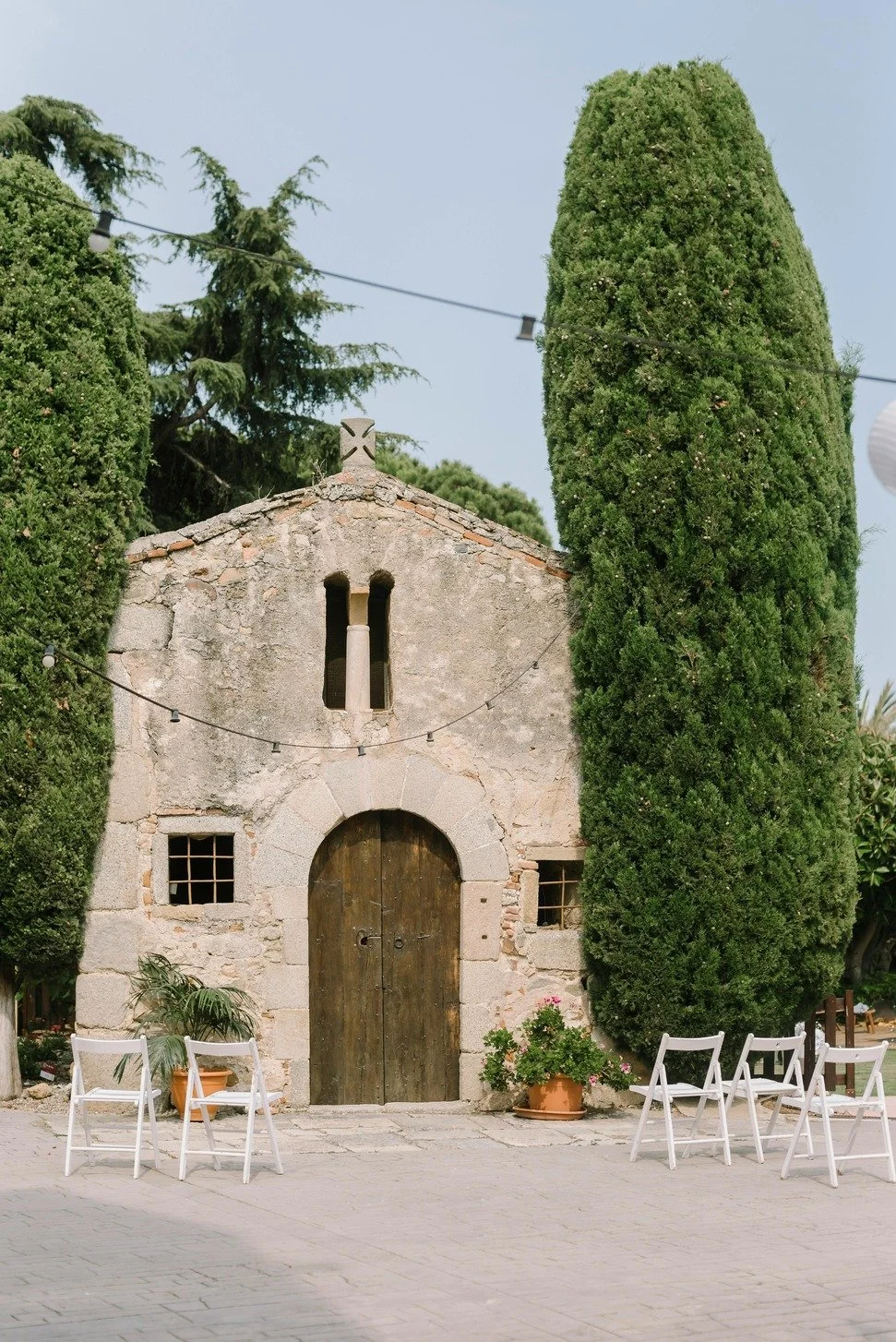 Small stone building with arched wooden door, two narrow windows, surrounded by tall green trees and potted plants, outdoor seating with white chairs, string lights overhead.