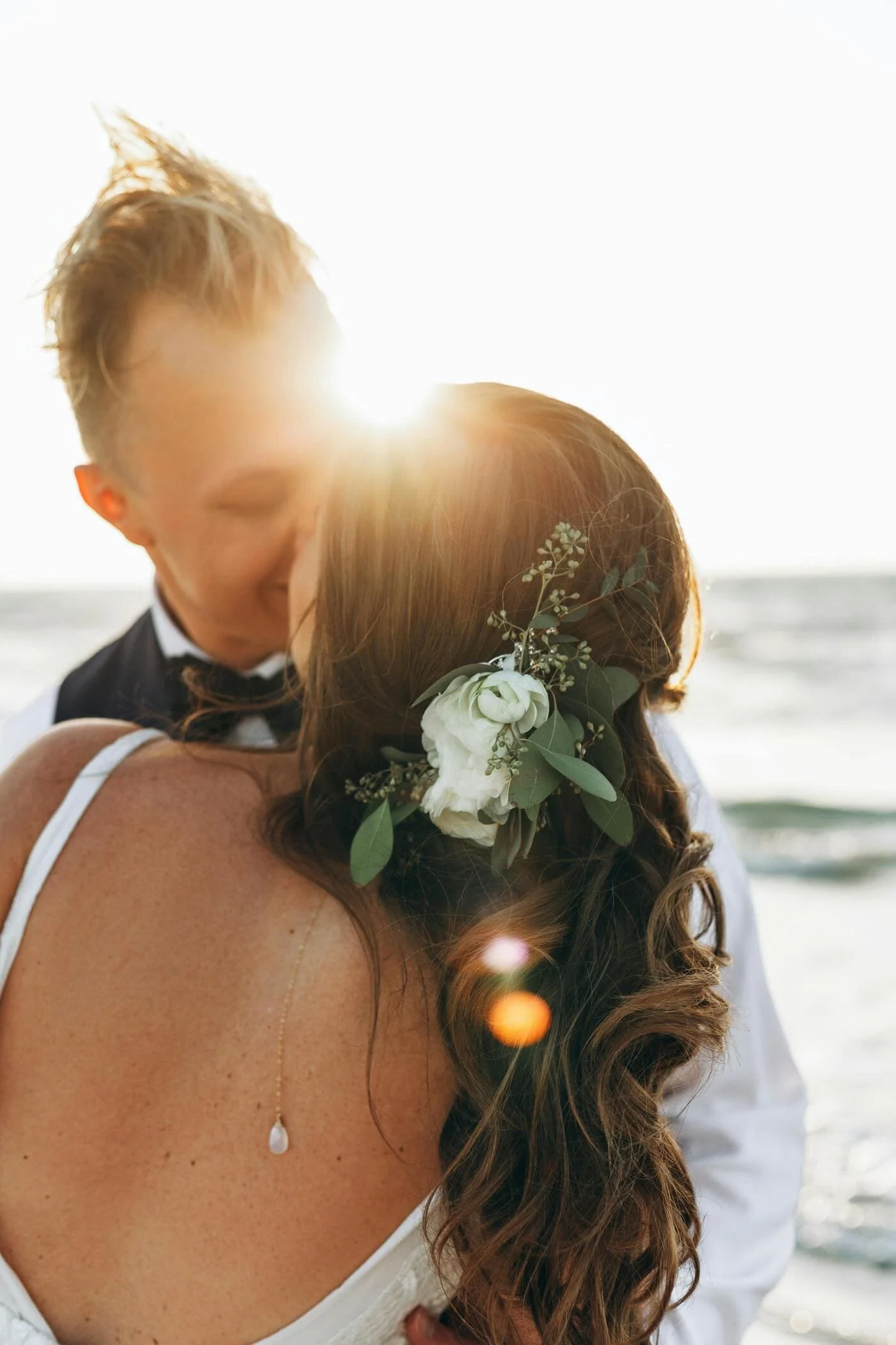 A couple hugging on the beach during sunset, with the sun behind them and ocean waves in the background. The woman has a floral hair accessory and is wearing a white dress with a delicate necklace. The man is dressed in a white shirt and dark vest.