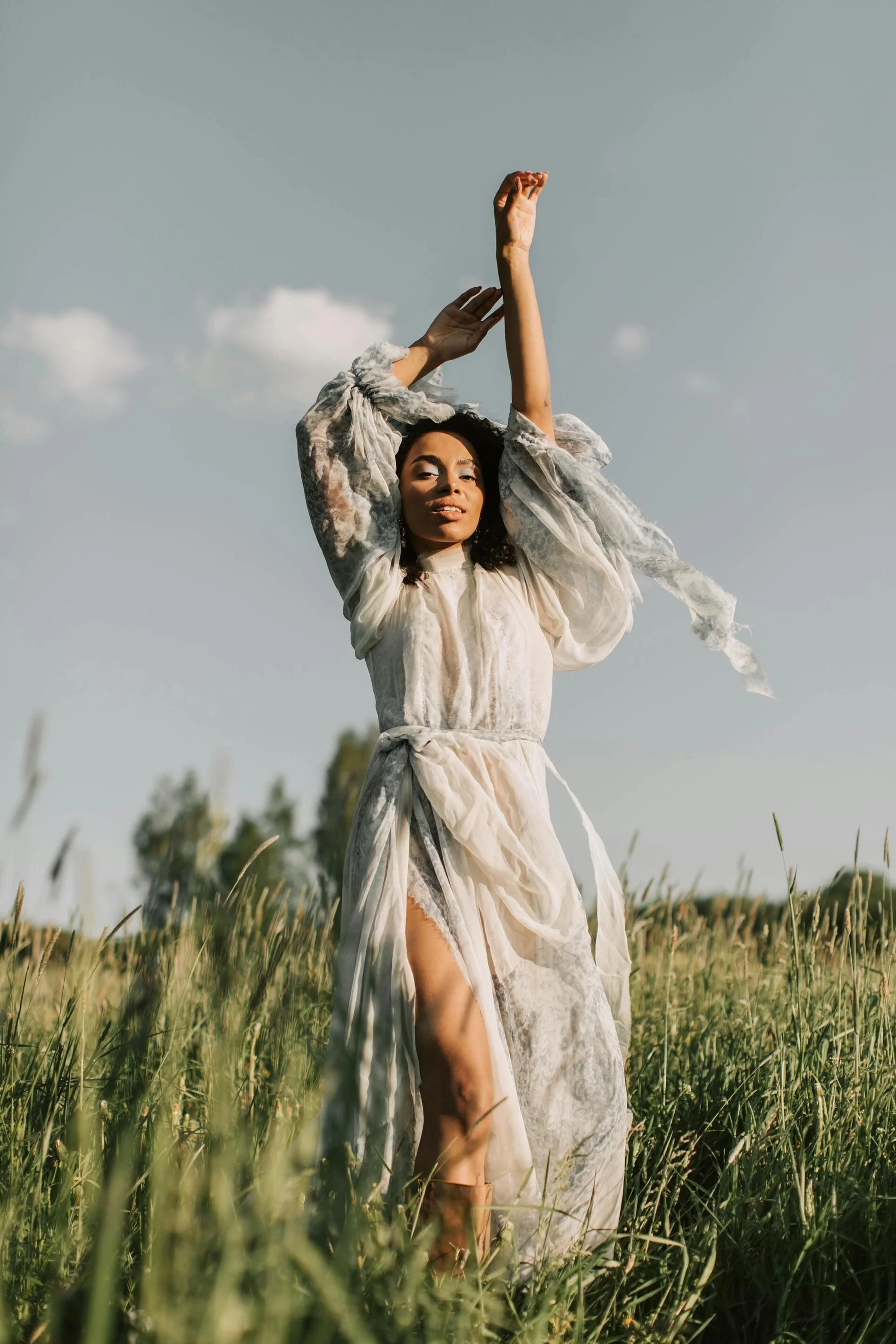 A woman in a flowing white dress standing in a grassy field with her arms raised above her head on a clear day.