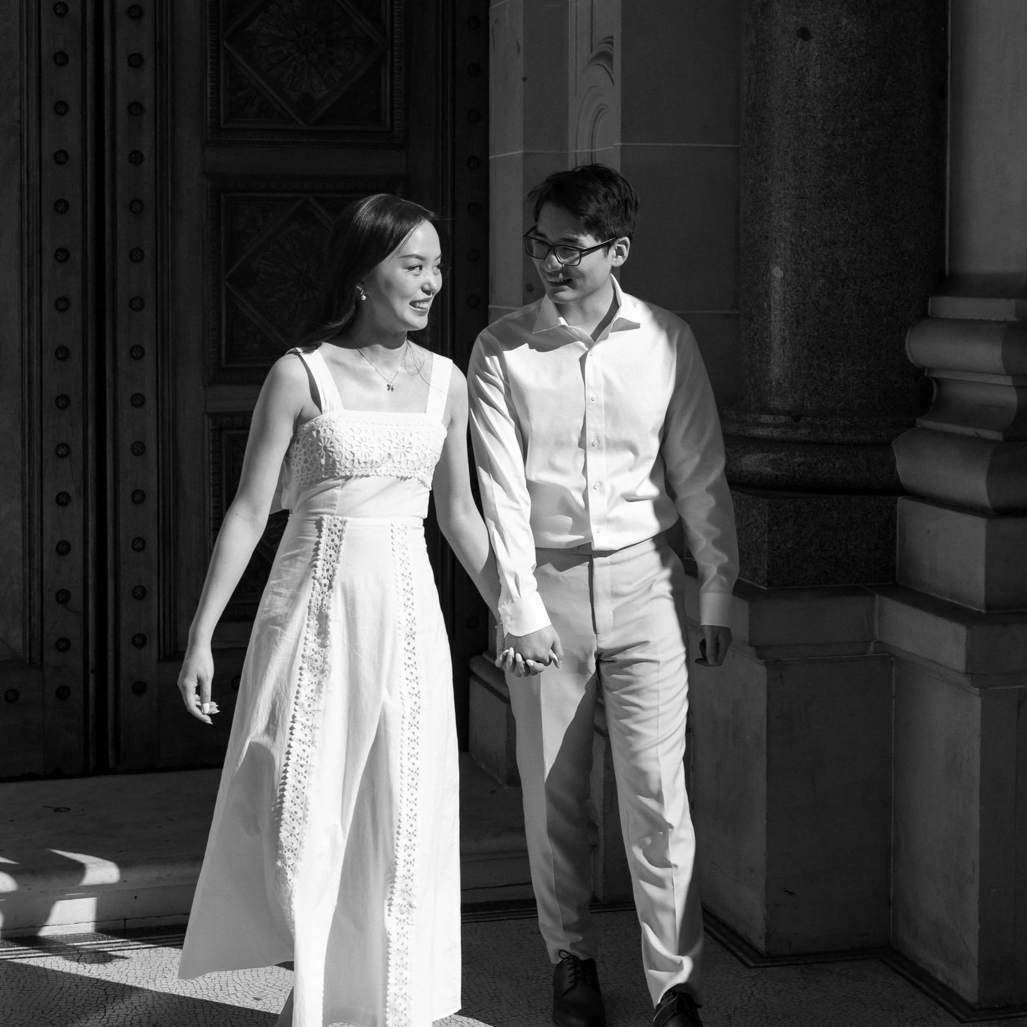 A black-and-white photo of a couple holding hands and walking out of a building with ornate wooden doors and stone columns.