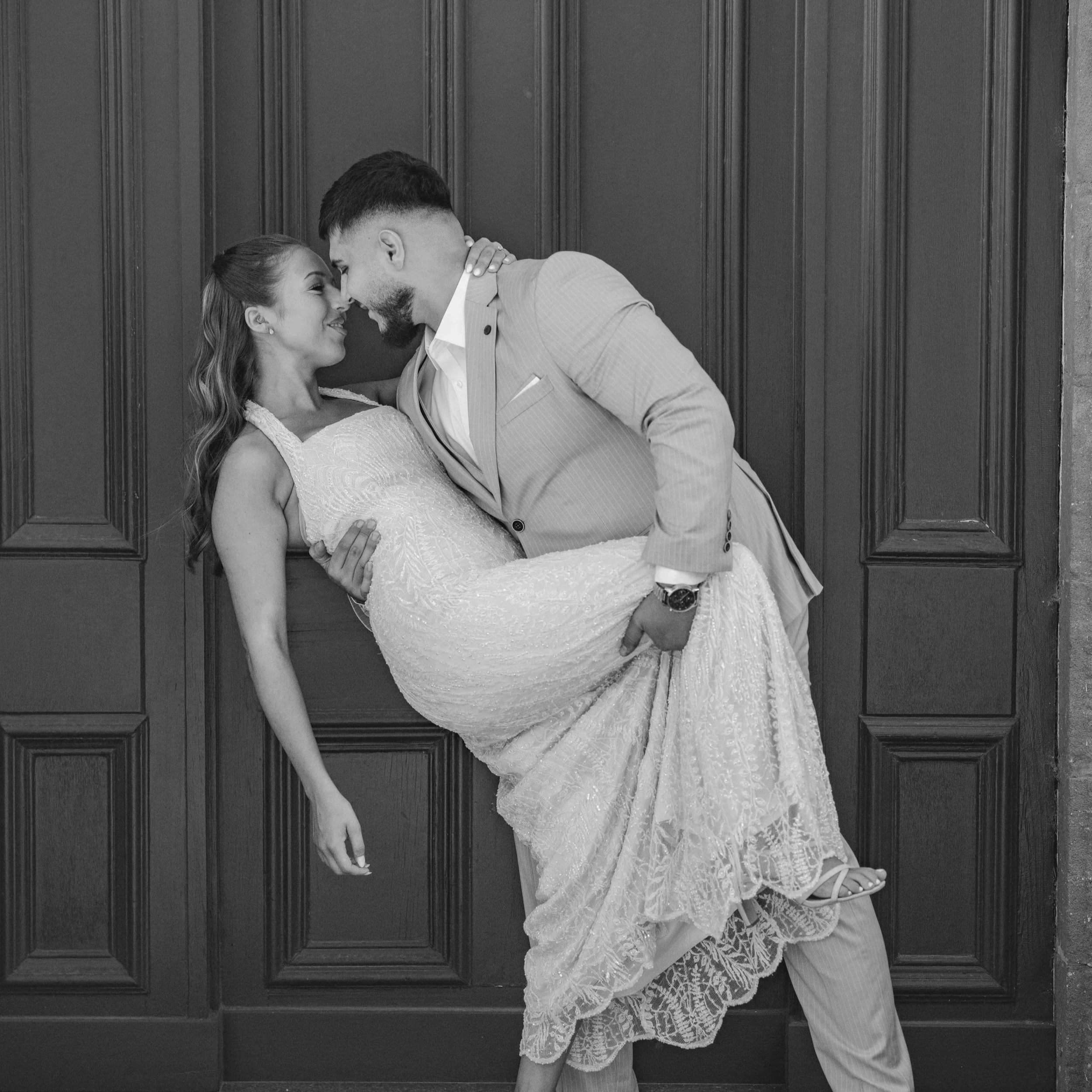 A couple dressed in wedding attire holding each other and leaning their foreheads together in front of wooden paneling.