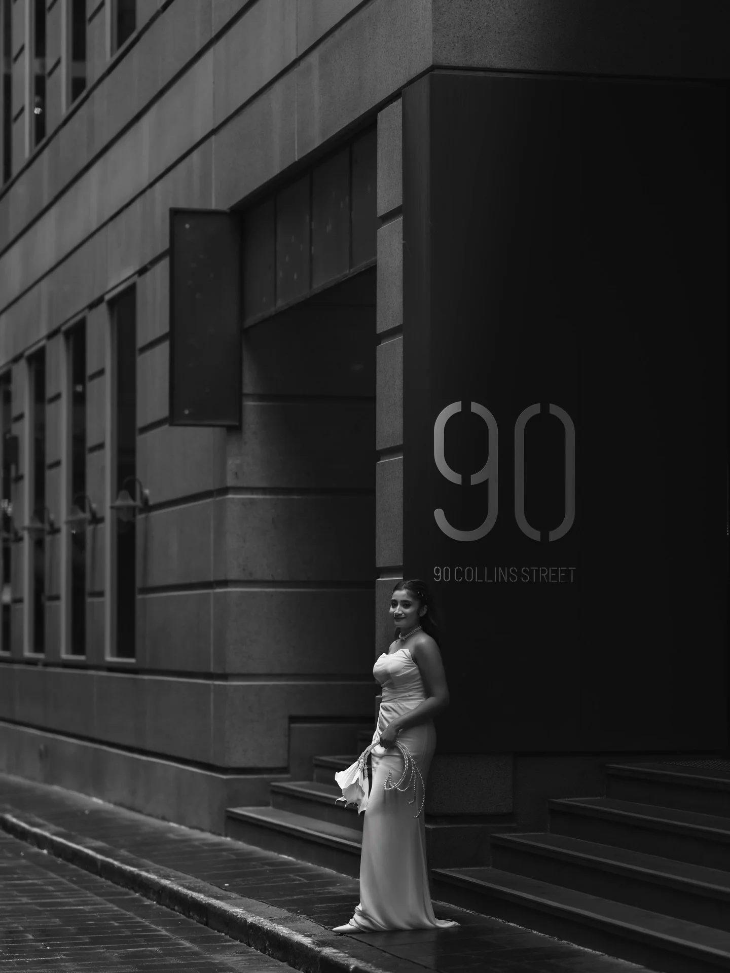 A woman wearing a strapless gown holding a purse stands on a city sidewalk in front of a large sign for 90 Collins Street, with an apartment building and windows in the background.