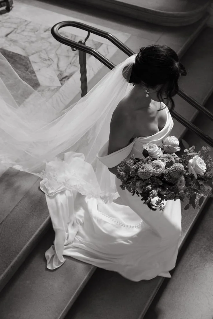 A bride sitting on stairs holding a bouquet of flowers, wearing a wedding dress and veil, with her head turned down.