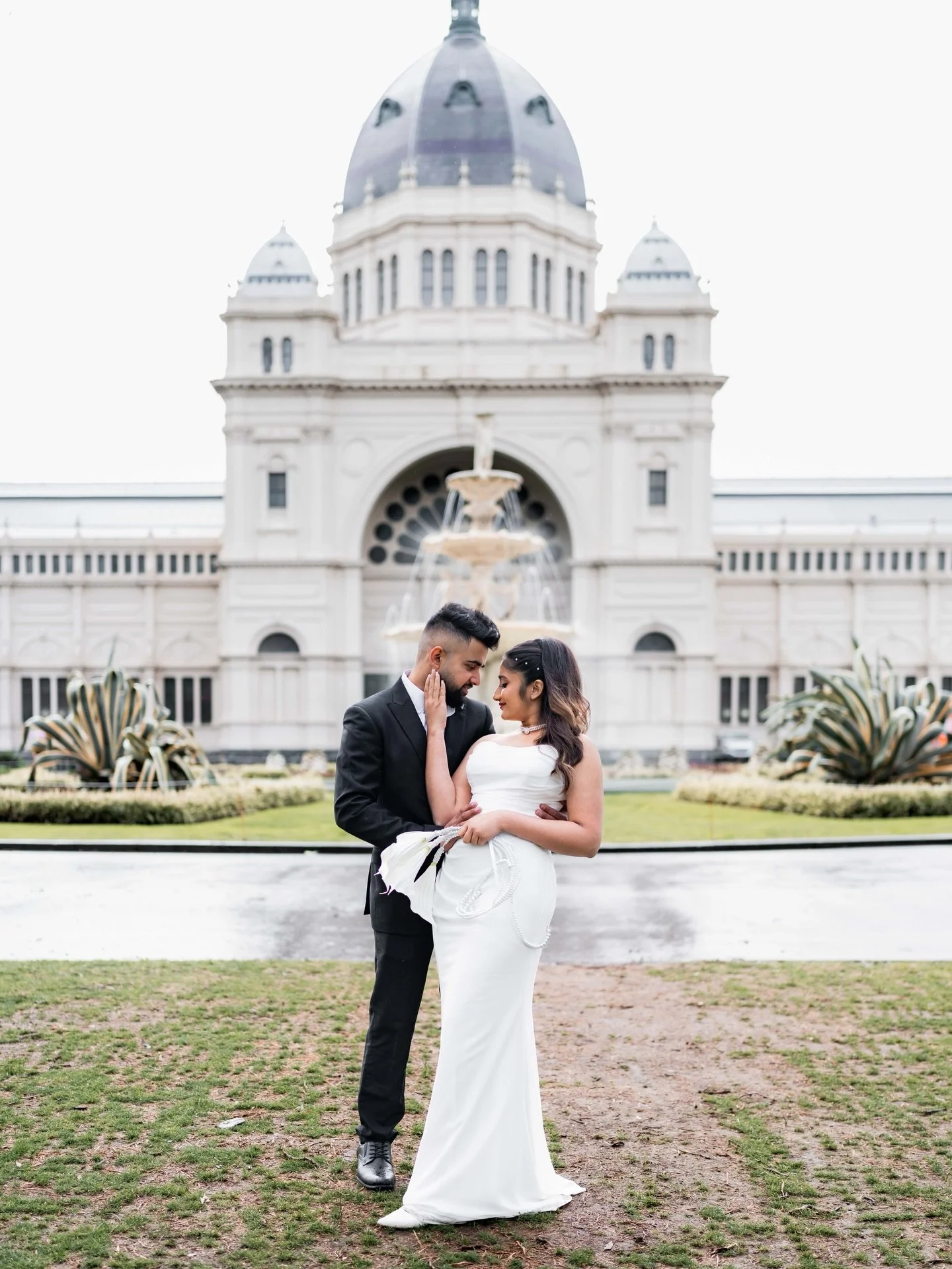 A couple in formal wedding attire standing close, with the man touching the woman's face, in front of a historic building with a fountain and formal landscaping.